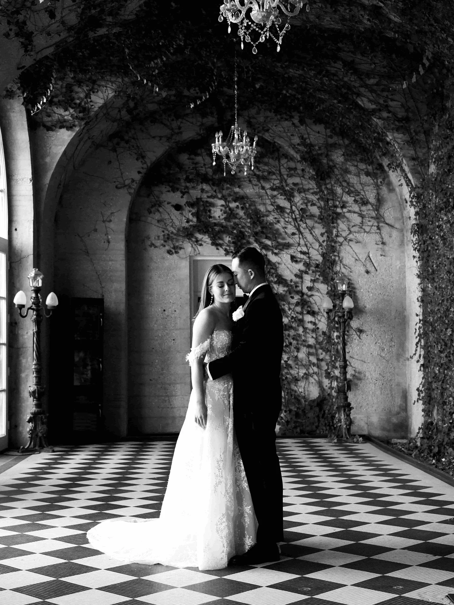 Black and white photograph of a bride and groom sharing a kiss in an elegant, arched indoor setting with chandeliers and patterned floor.