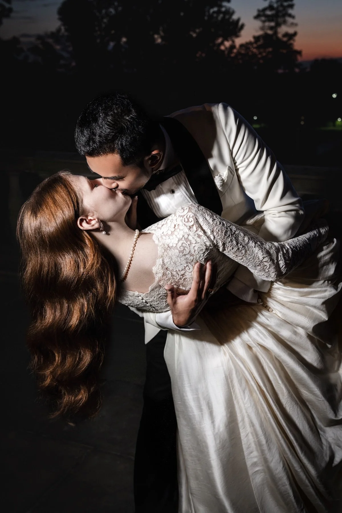 A couple dressed in formal wedding attire sharing a kiss outdoors during sunset or twilight.