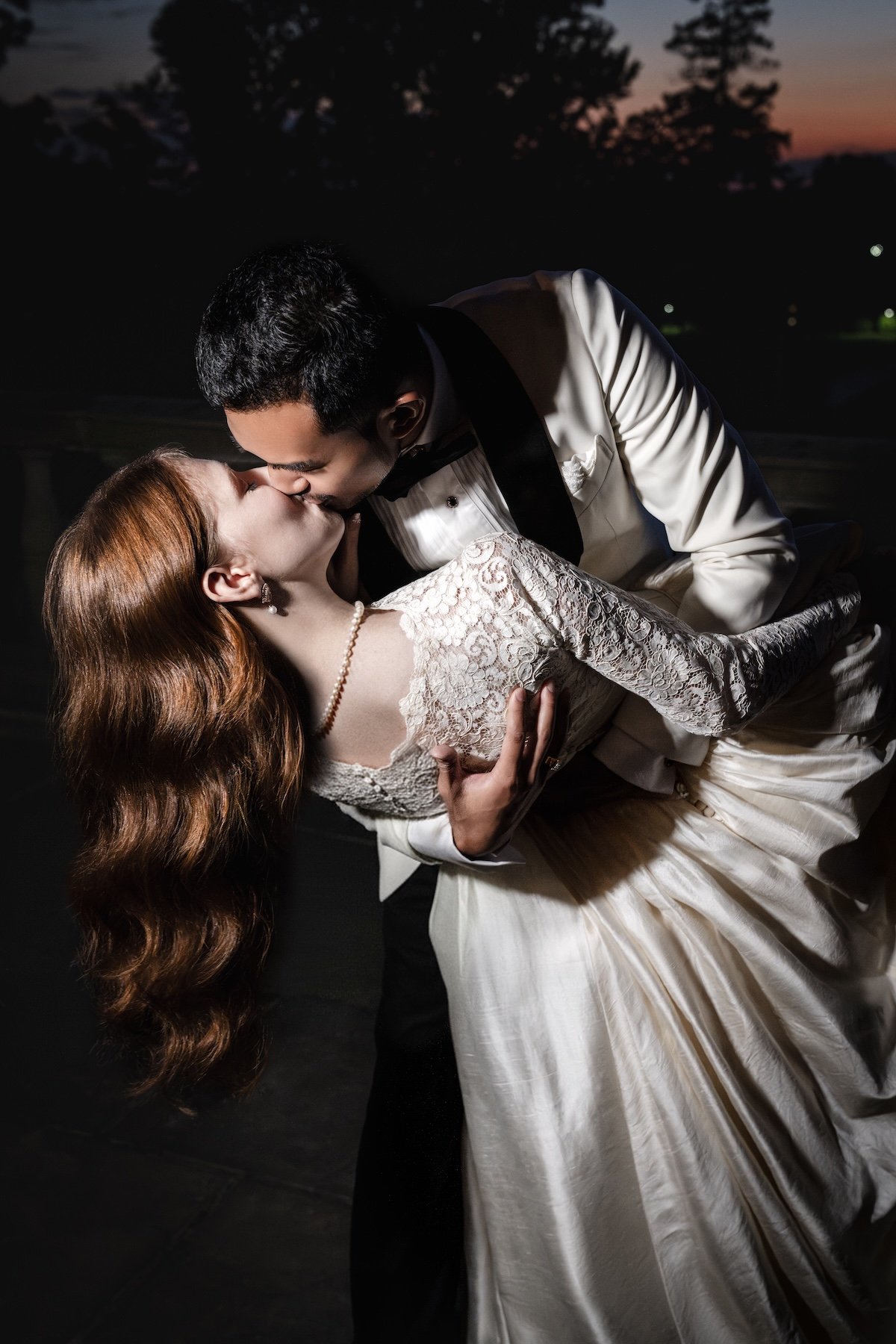 A couple sharing a romantic kiss outdoors during sunset, with the man dressed in a tuxedo and the woman in a lace wedding dress.