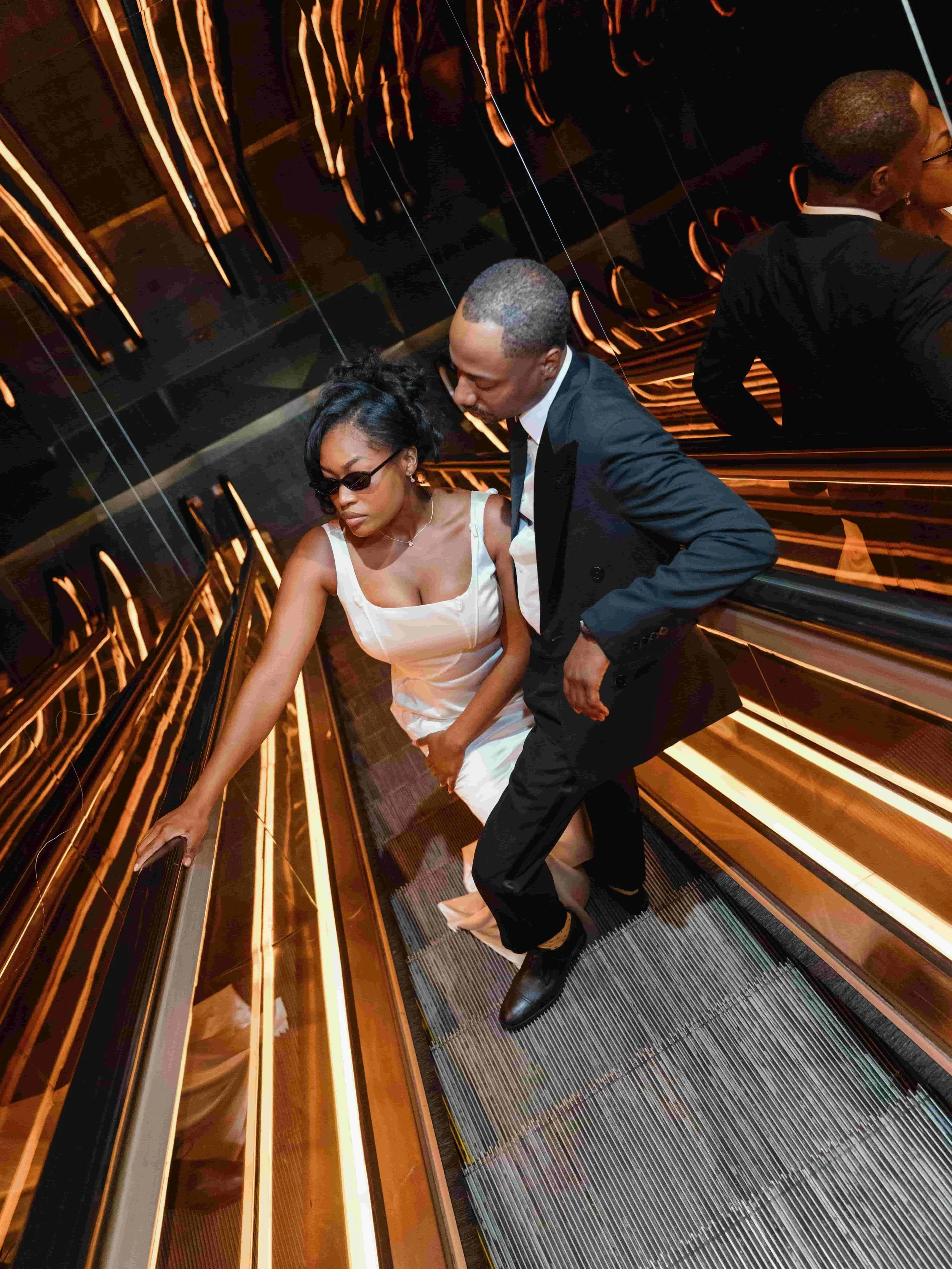 A bride in a white dress and sunglasses, and a man in a black suit and white shirt, stand on an escalator with a reflective gold and black wall behind them.