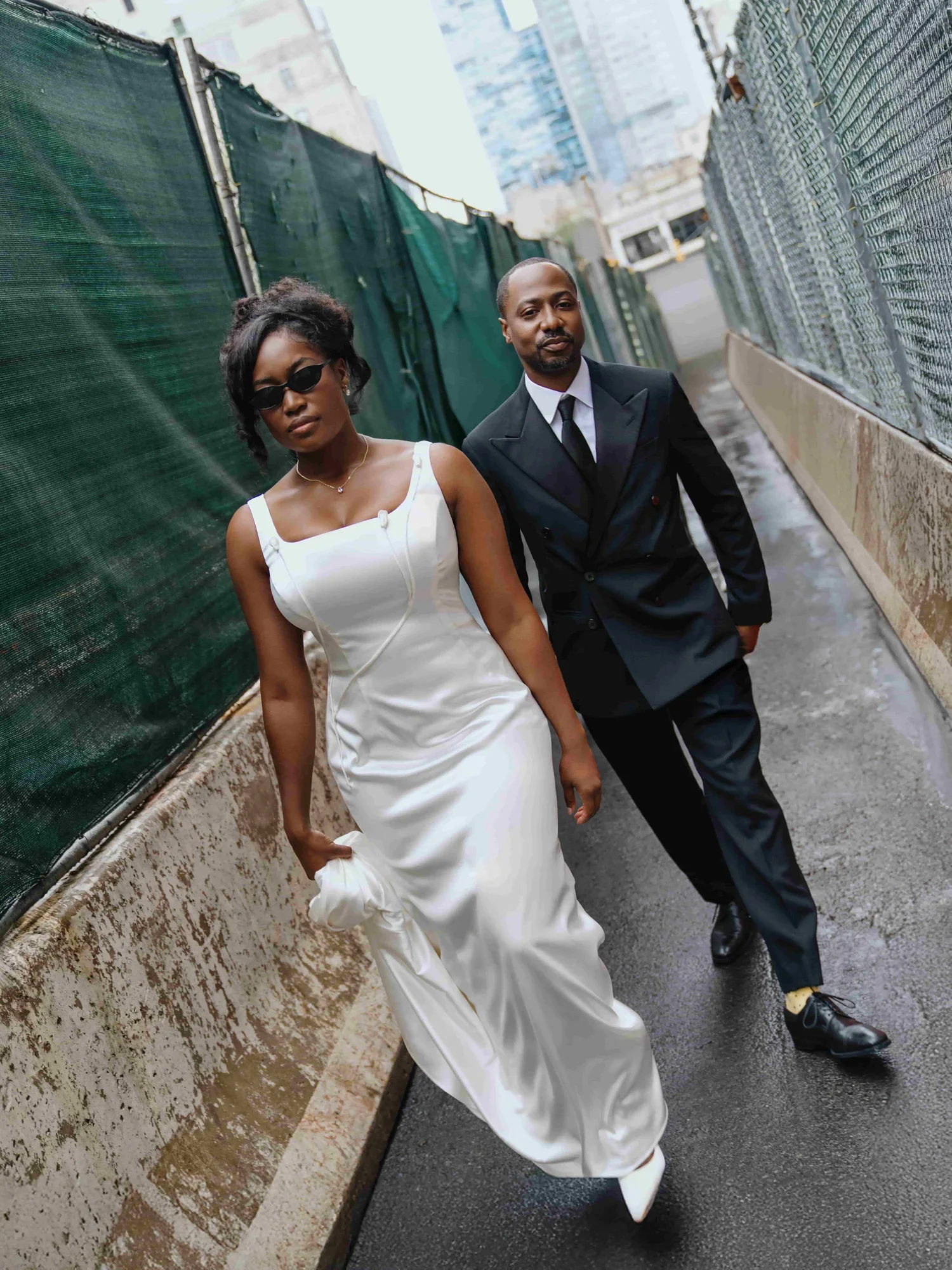 A stylish couple walking outside in an urban setting, dressed in formal attire with the woman in a white gown and the man in a black suit.