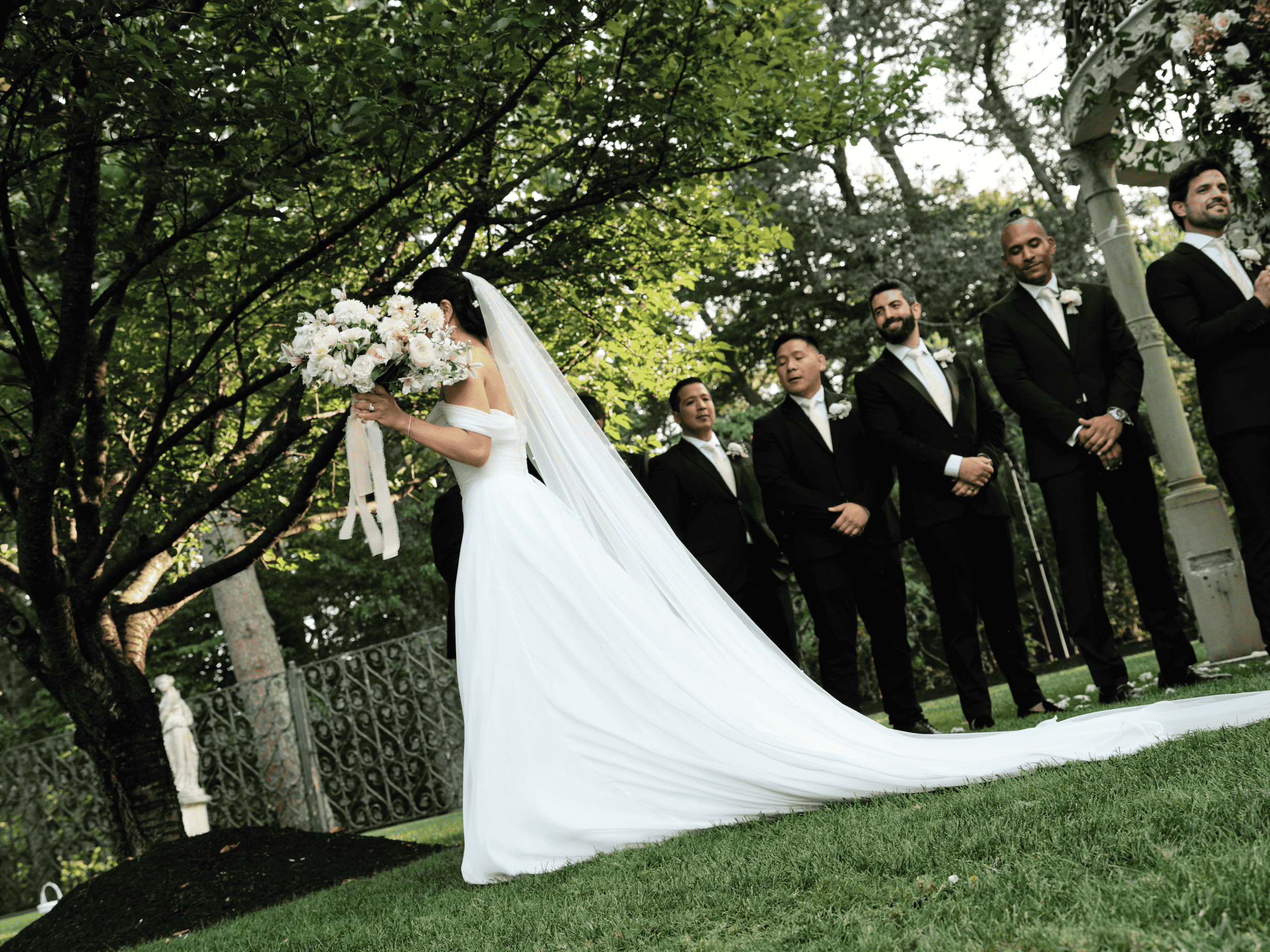 Bride holding a bouquet of white flowers during a wedding ceremony outdoors with seven men in black suits standing behind her.