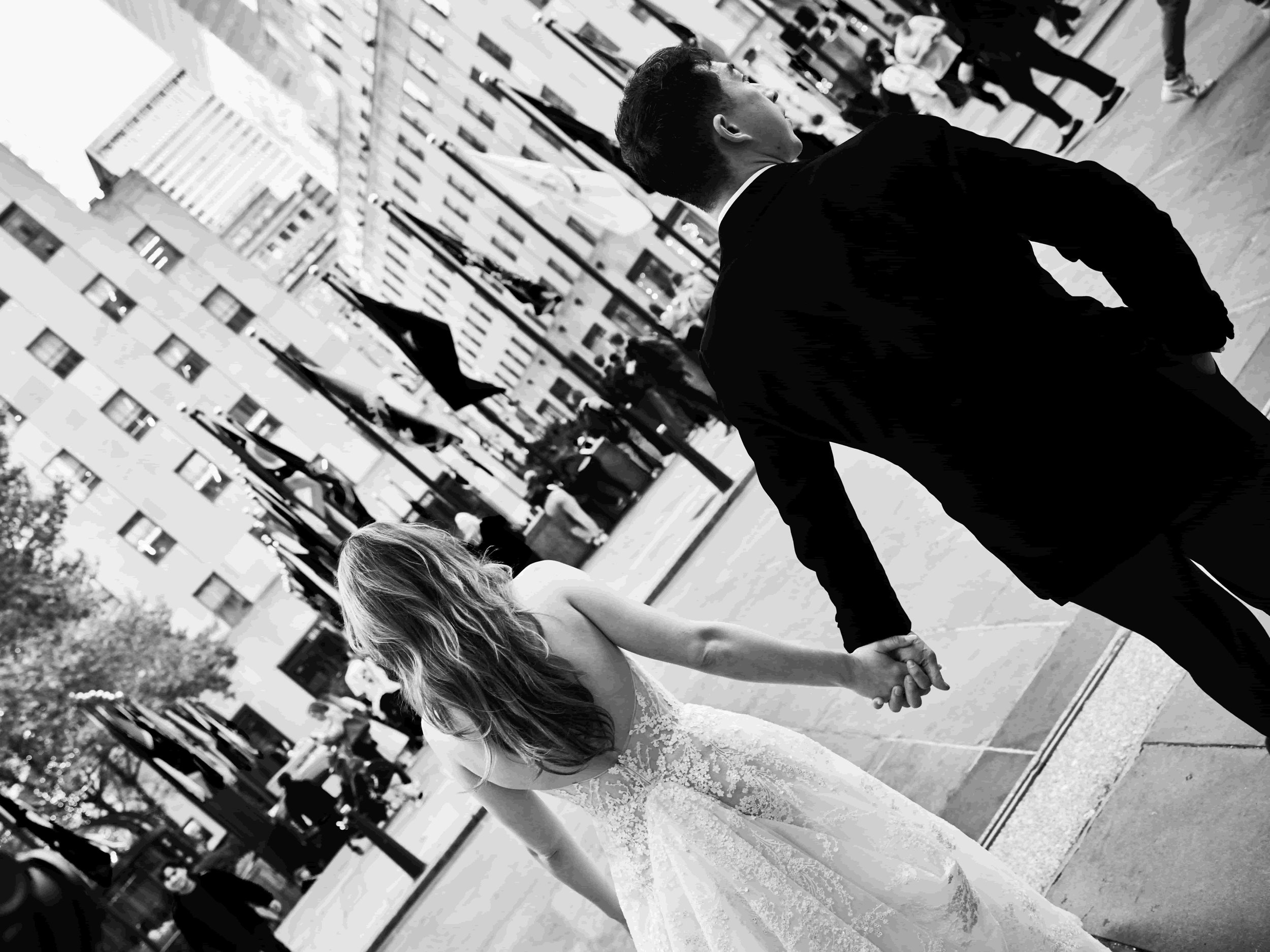 A bride and groom holding hands on a city sidewalk, with a crowd and tall buildings in the background, black and white photo.