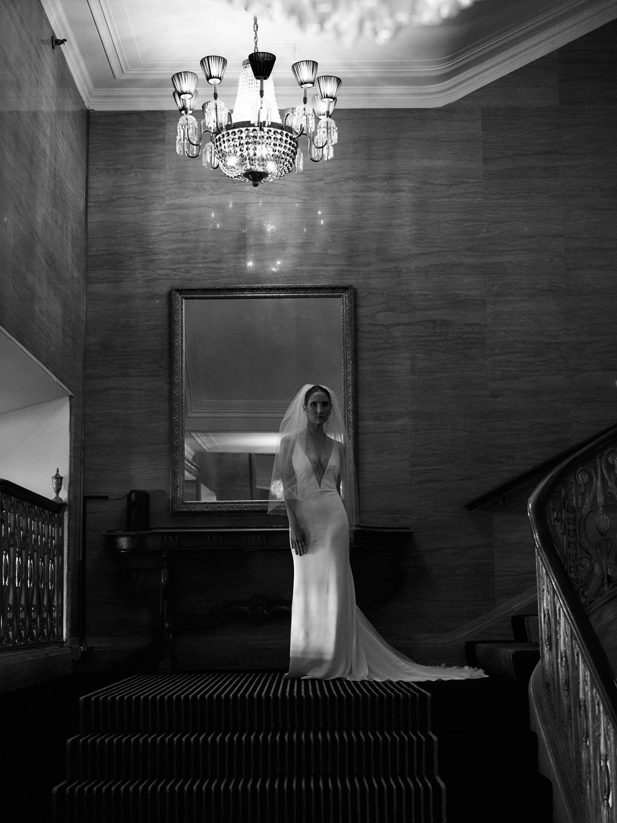 A bride in a long white wedding dress with a veil stands on a staircase landing in front of a large mirror in a luxurious, dimly lit room with wood-paneled walls and an ornate chandelier hanging from the ceiling.