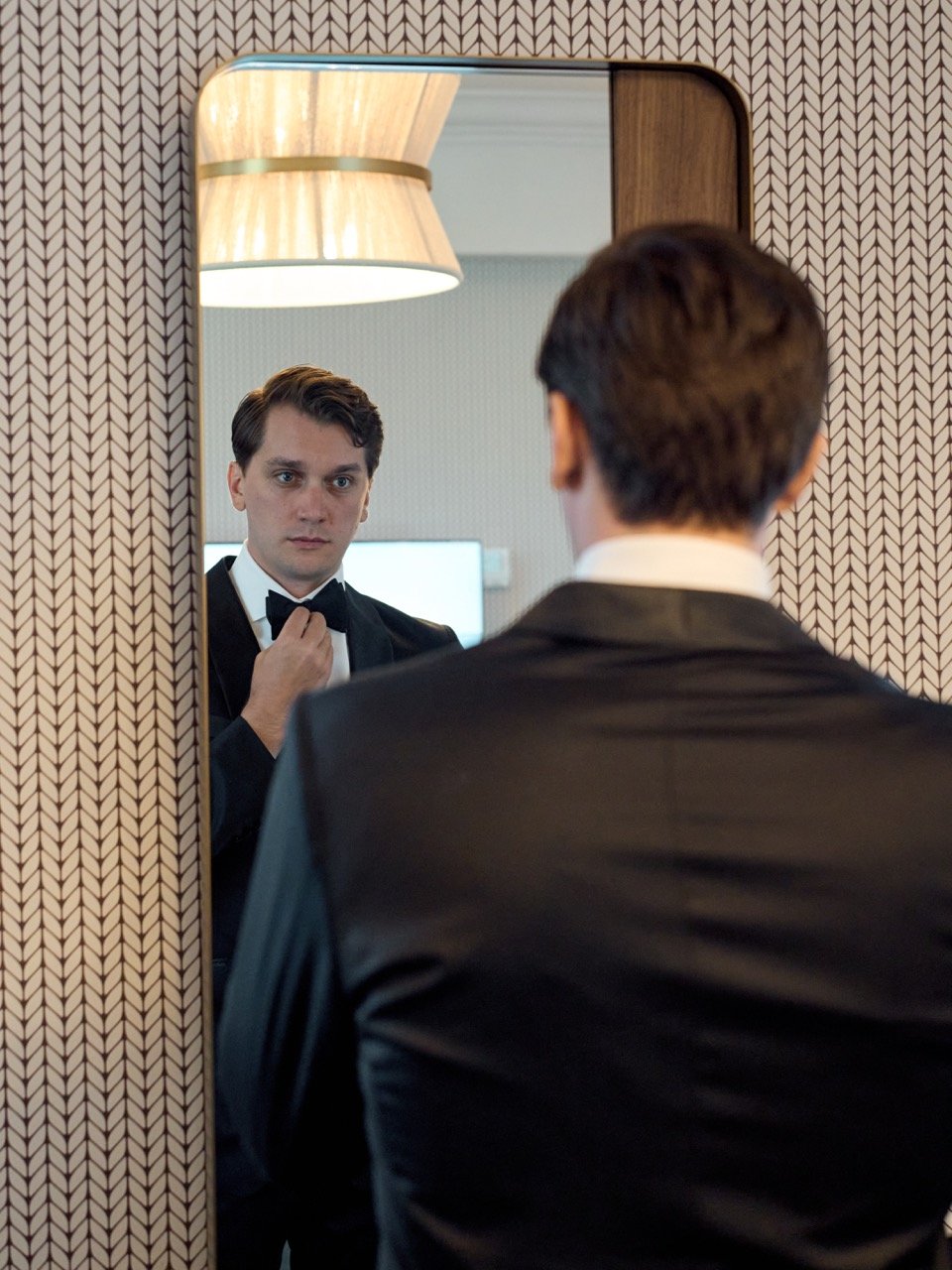 A man in a tuxedo adjusting his bow tie while looking into a mirror, with patterned wallpaper and a hanging lamp in the background.