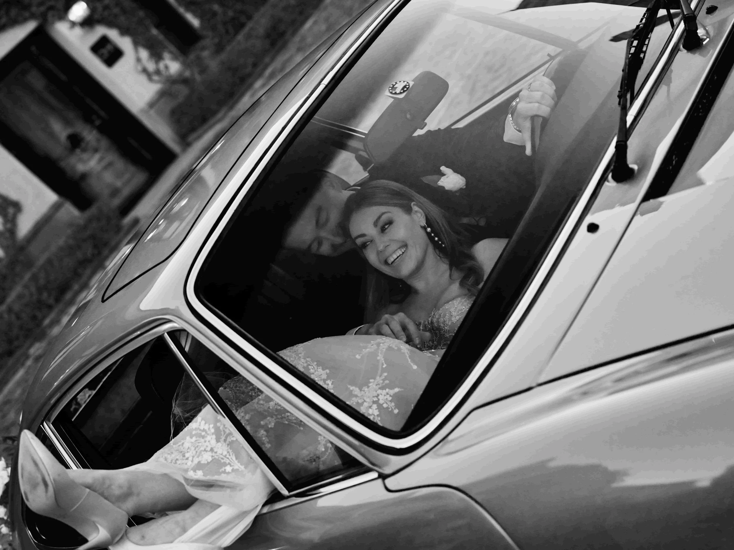 Black and white photo of a bride and groom sitting inside a vintage car, smiling and holding hands, with bride showing her wedding dress and high heels through the window.