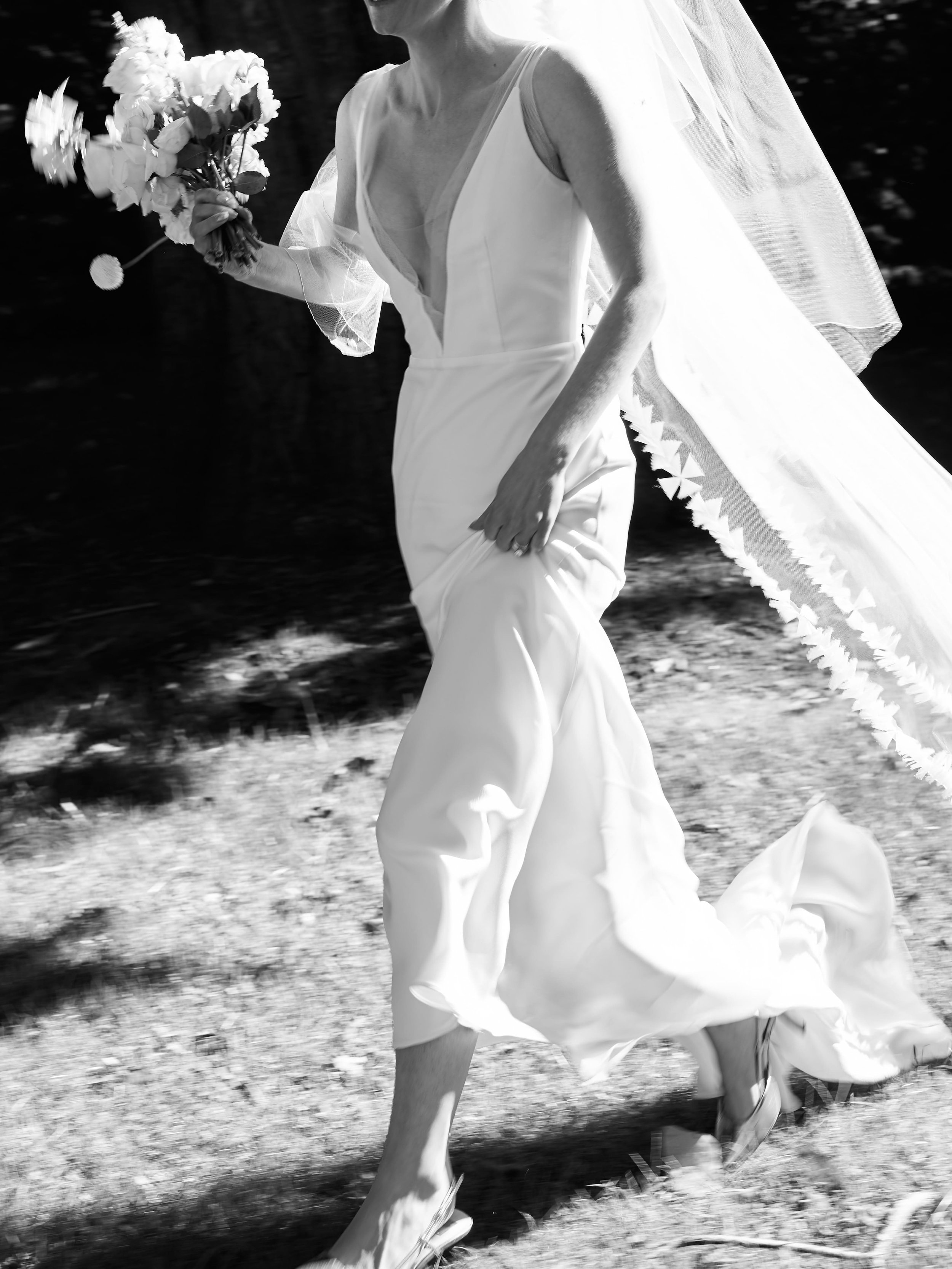 Black and white photo of a woman in a wedding dress holding a bouquet, walking outdoors.