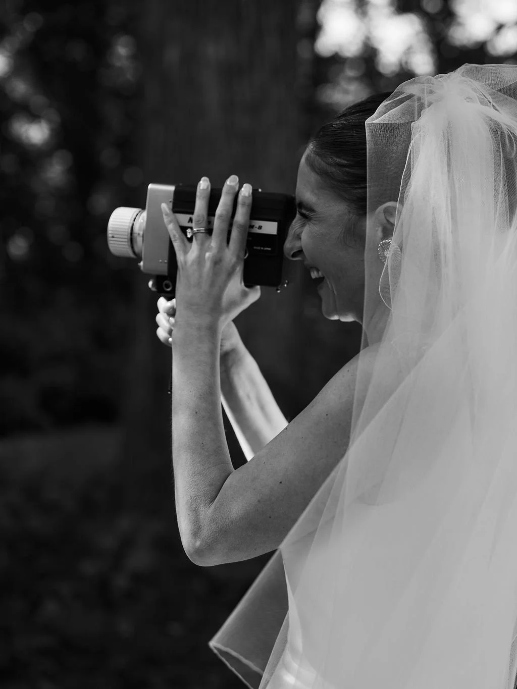 Black and white photo of a smiling bride wearing a veil, holding a vintage camera to her eye, outdoors.