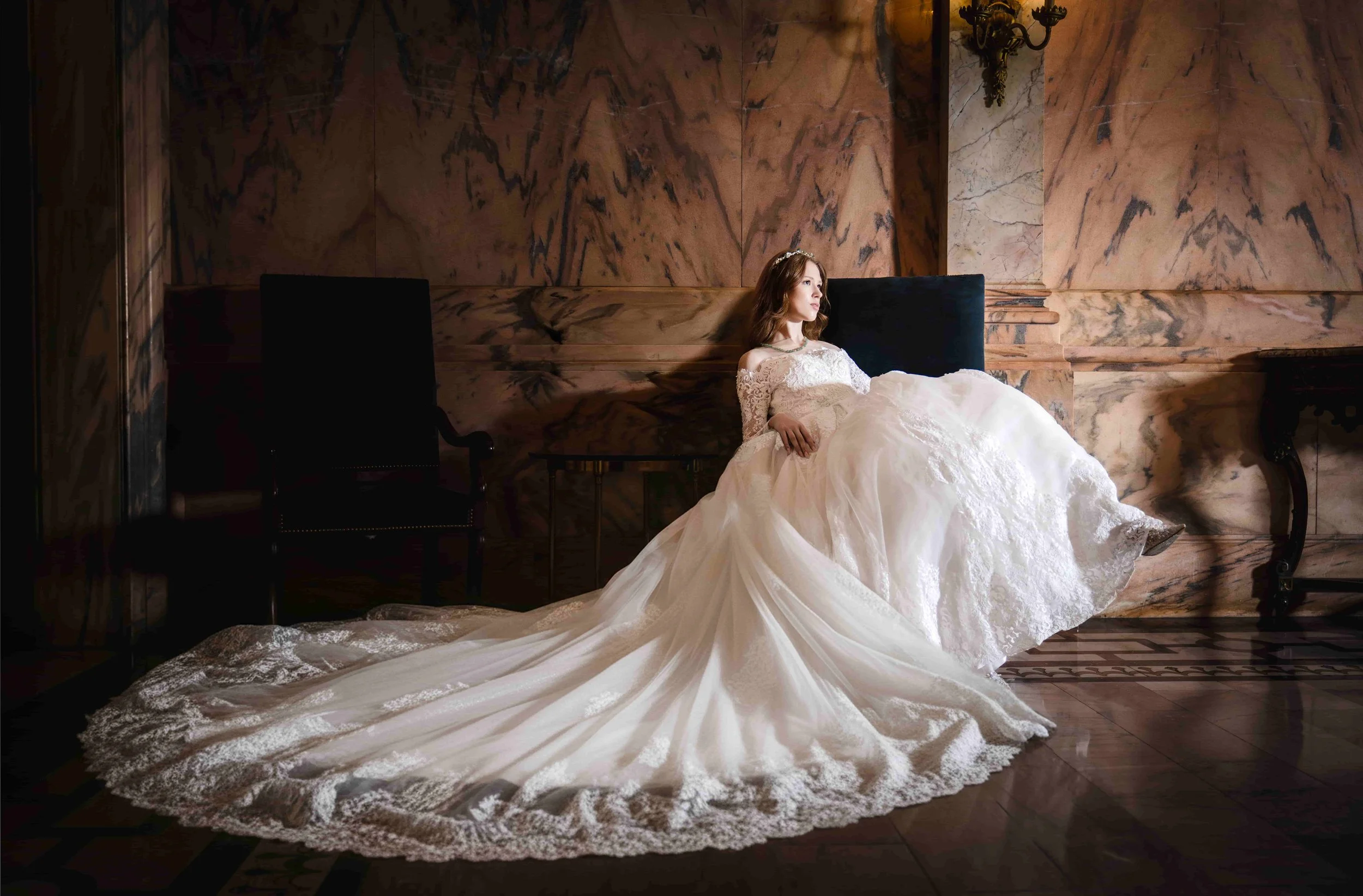 A bride in a white wedding gown sits on a cushion, leaning against a dark headboard, in a dimly lit ornate room with marble walls and antique furniture.