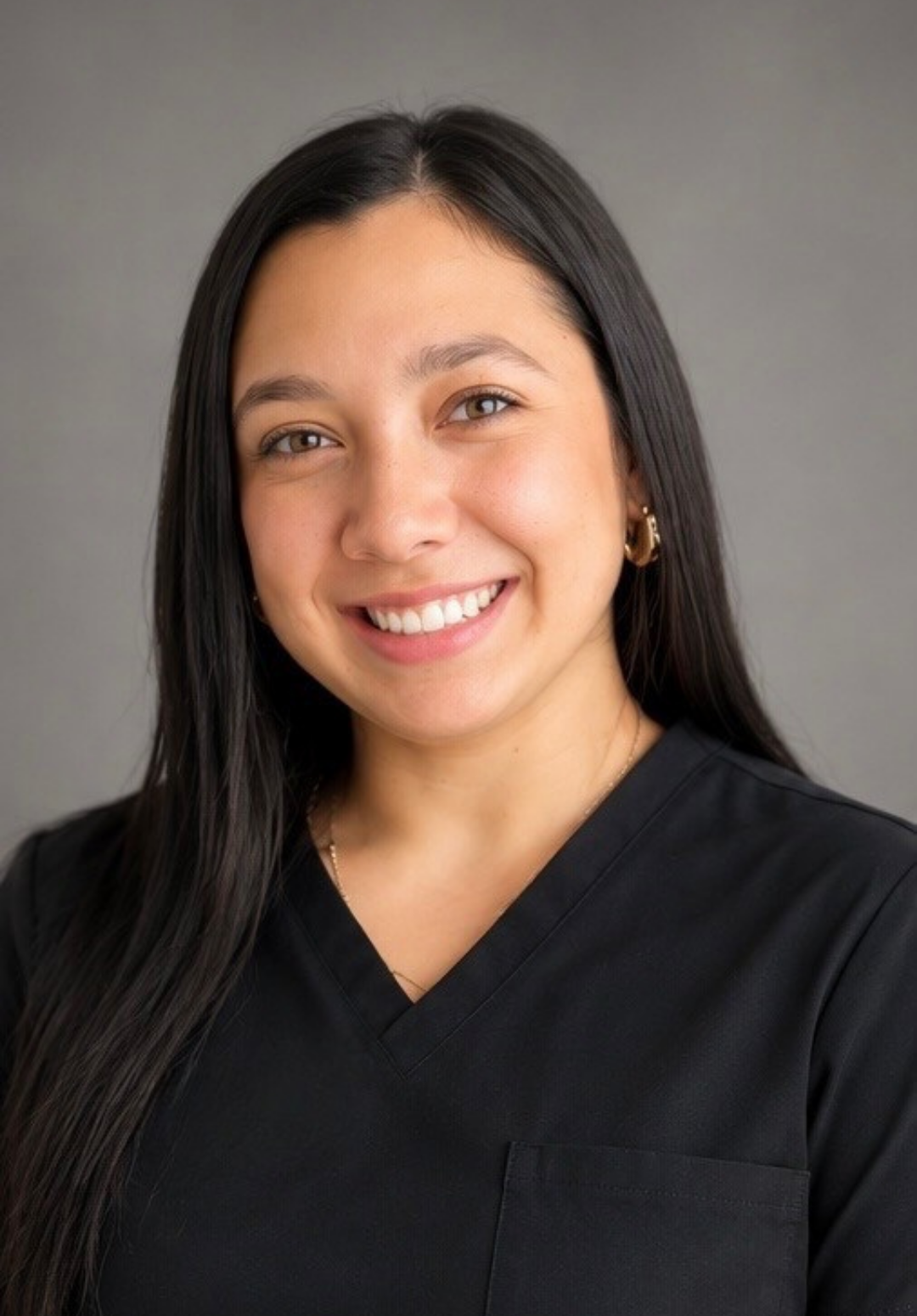 A woman with long dark hair smiling wearing medical scrubs and a name tag, standing against a plain light-colored wall.