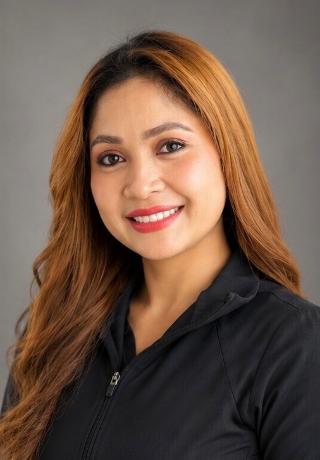 A woman with long curly brown hair and a bright smile, wearing a black jacket with the logo of Rosemarie Dental on it, standing against a plain off-white wall.