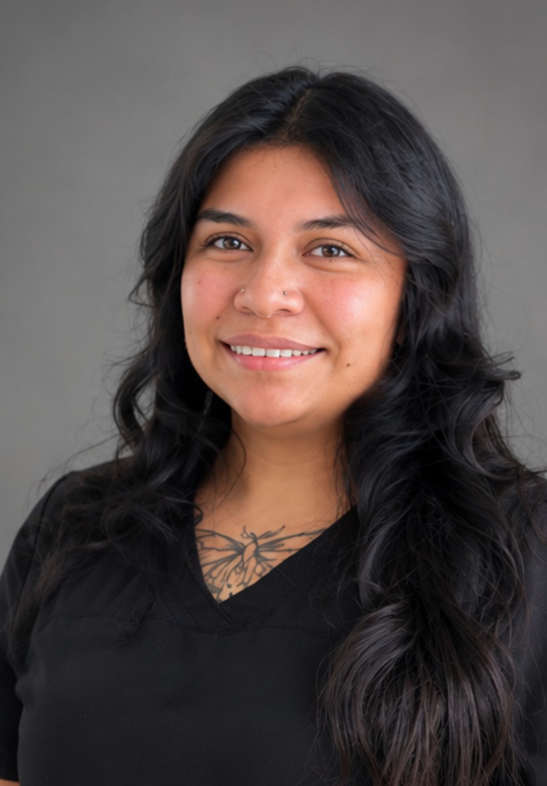 Portrait of a woman with long dark hair wearing a black uniform with a name tag, standing against a plain light-colored wall.