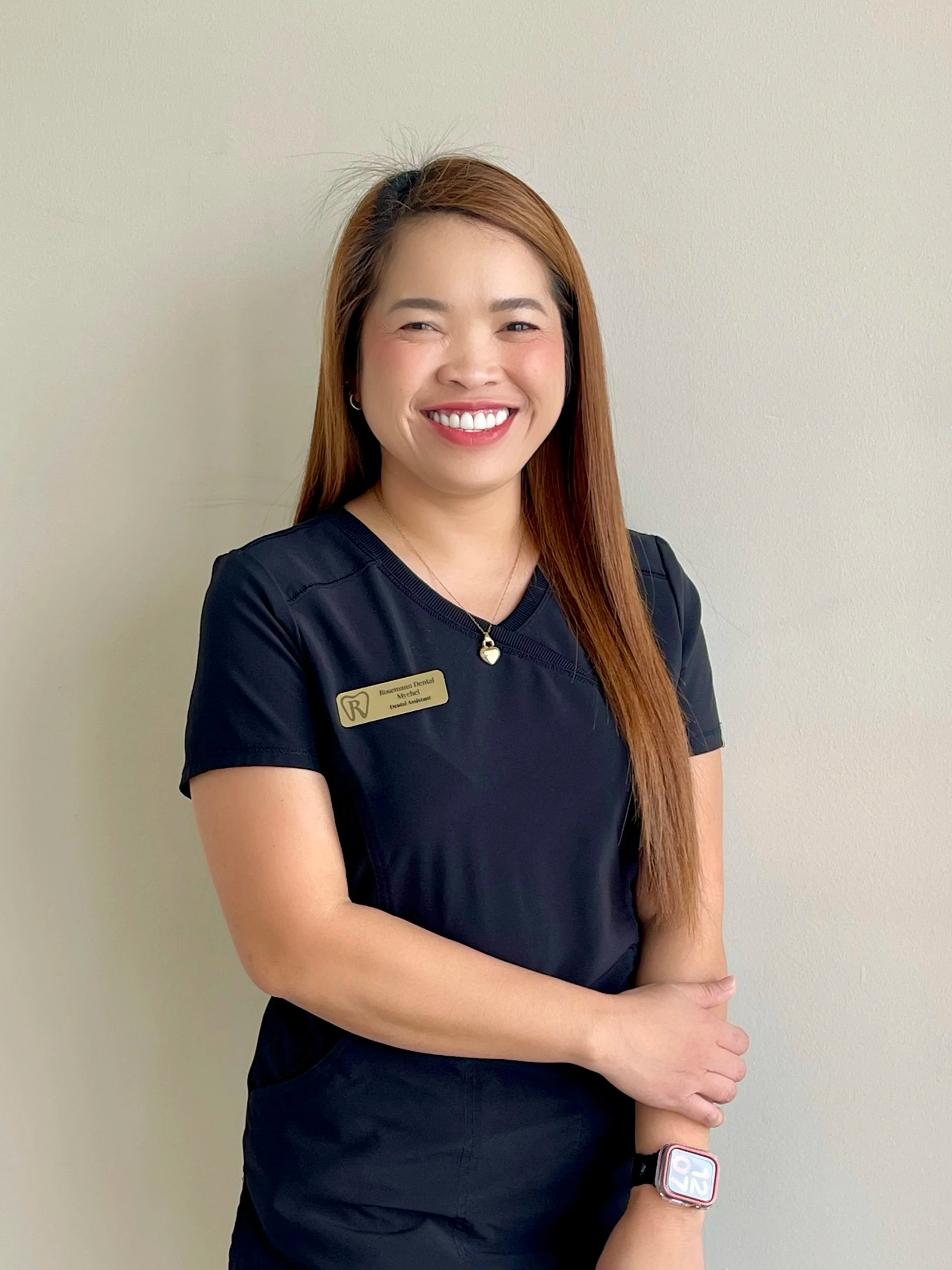 A woman with long dark hair smiling, wearing a black dental smock with 'Rosemann Dental' logo, standing against a plain wall.