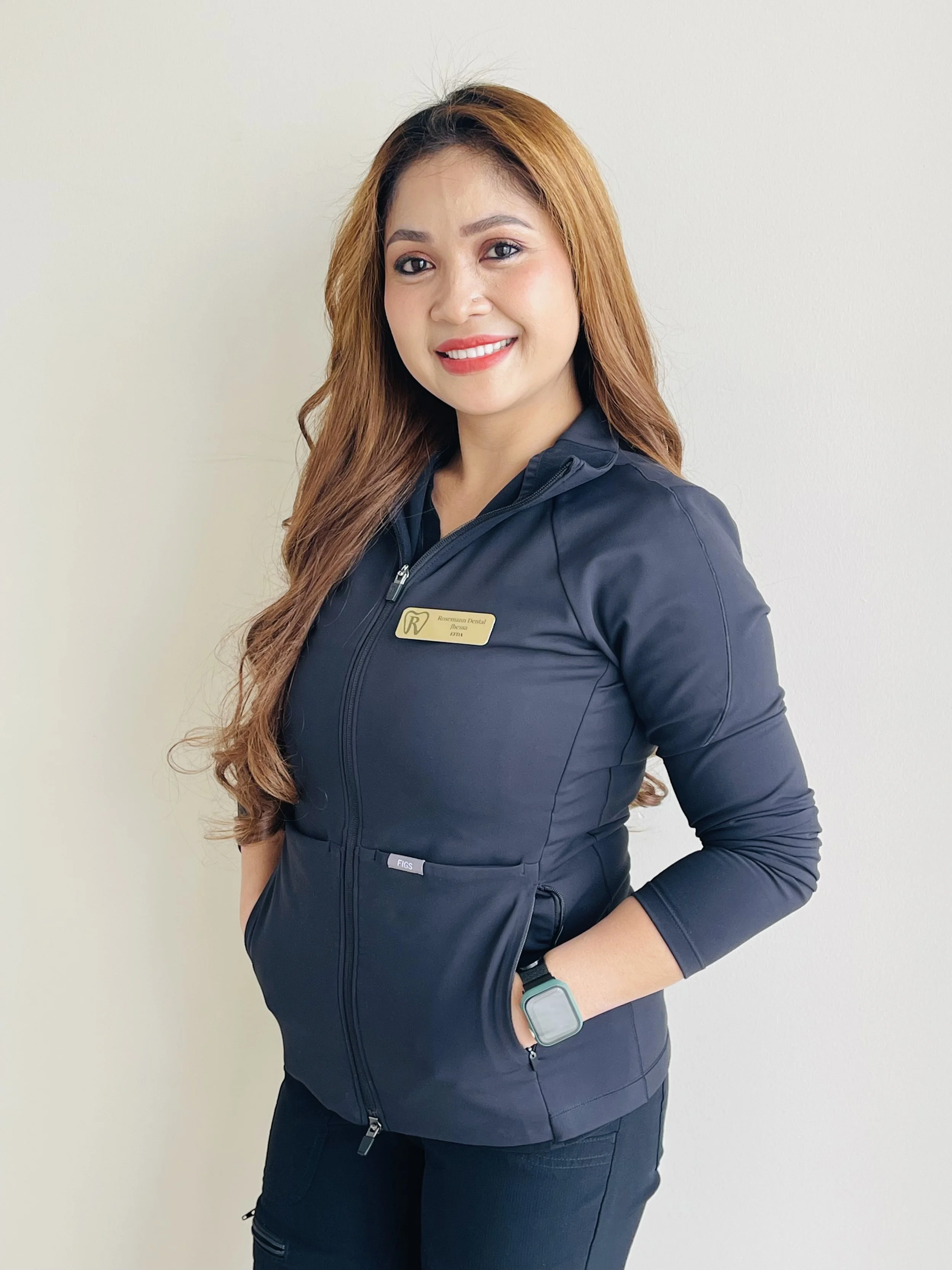 A woman with long curly brown hair and a bright smile, wearing a black jacket with the logo of Rosemarie Dental on it, standing against a plain off-white wall.