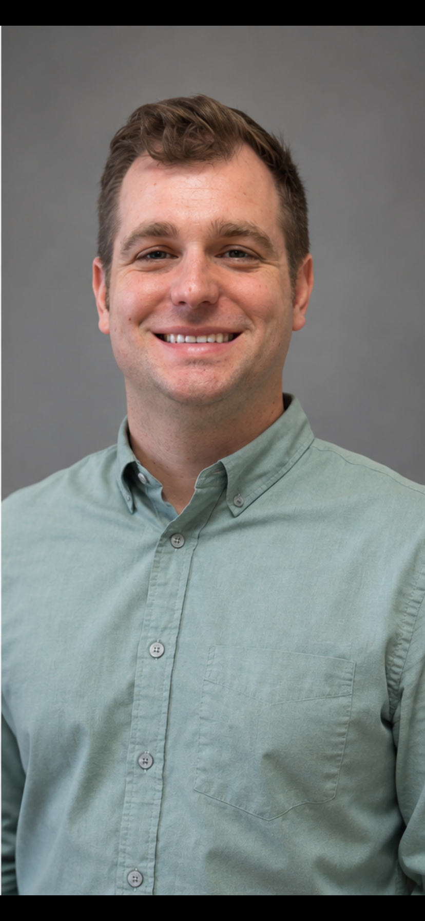 A man with brown hair and a light green button-up shirt, smiling and standing against a plain off-white wall.