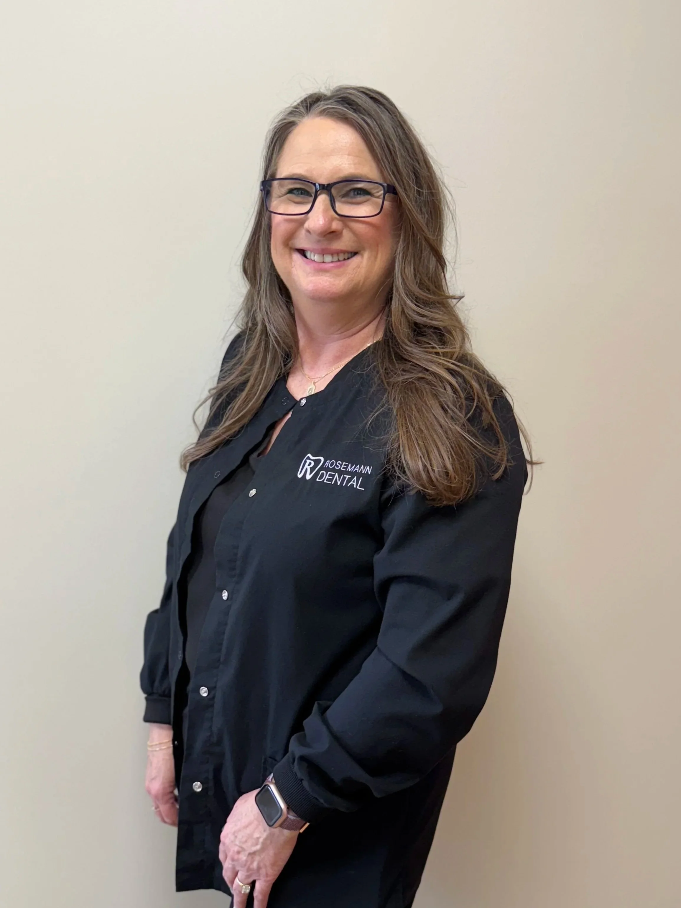 A smiling woman with long wavy brown hair and glasses wearing a black jacket with the Rosemann Dental logo, standing against a plain light beige wall.