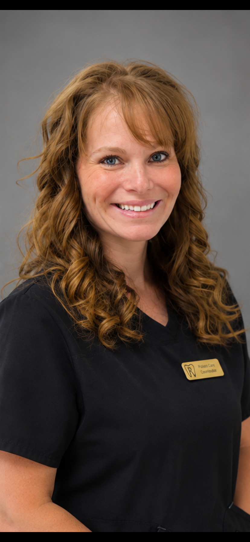 A woman with curly, strawberry blonde hair smiling and wearing a black jacket with the logo of Roseman Dental, standing against a plain light-colored wall.