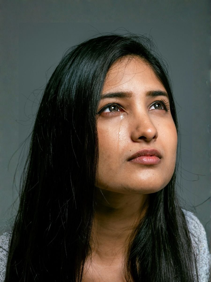 A young woman with long black hair looking upward, with tears on her face, against a gray background.