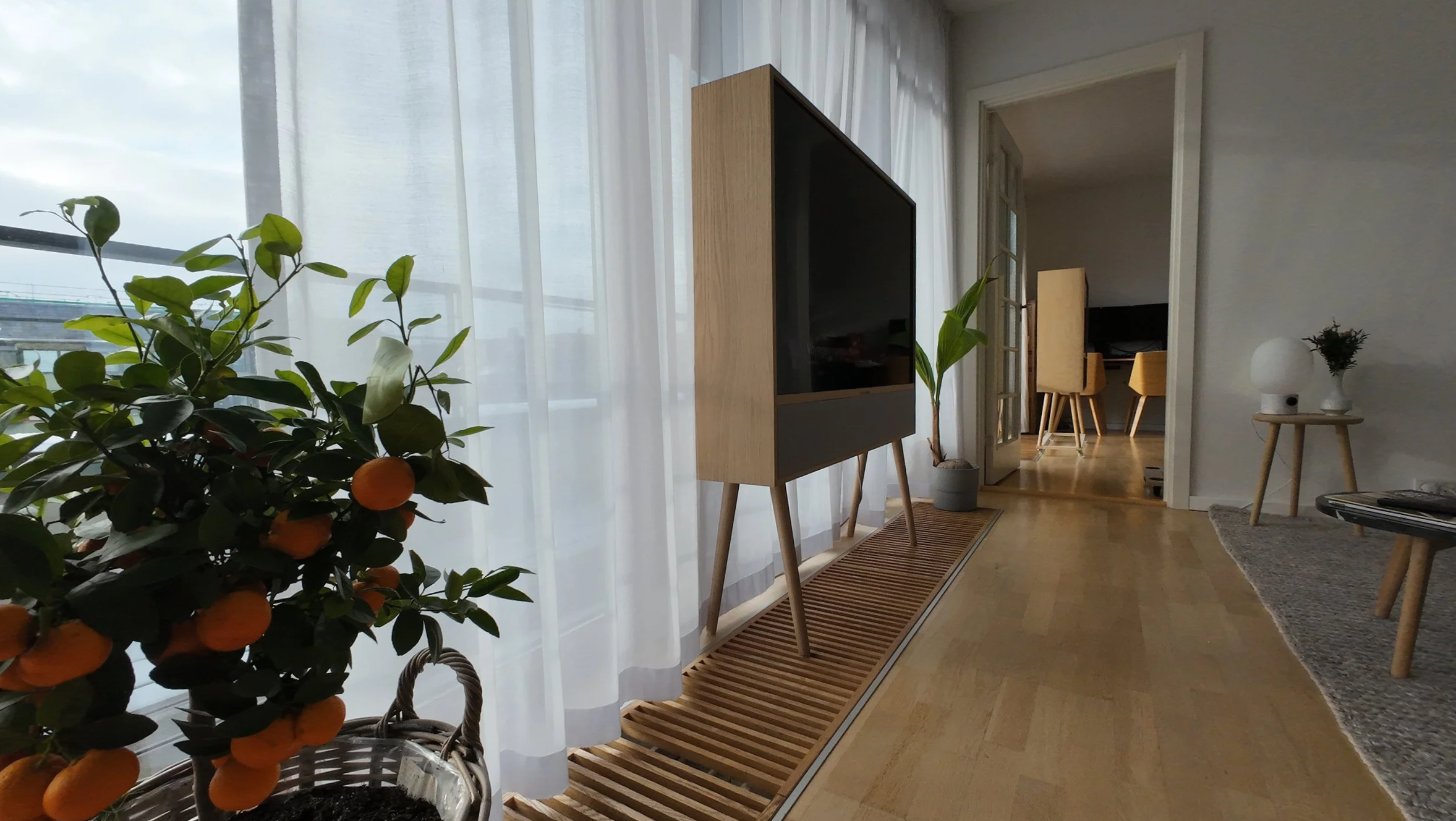 Living room with a wooden floor, potted plant with orange fruit near window, modern TV unit, and a doorway leading to a dining area with yellow chairs.