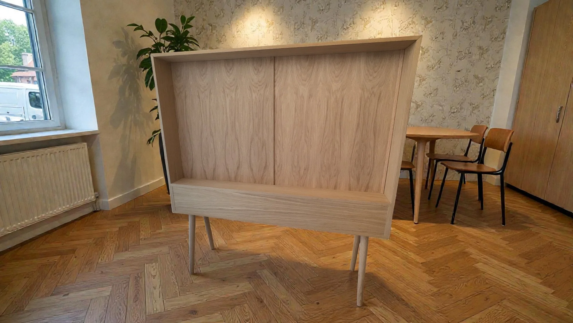Unfinished wooden bookshelf with a light natural finish, elevated on four angled legs, positioned in a room with herringbone parquet flooring, next to a window with a radiator below, a green plant nearby, and a round table with three chairs in the background.