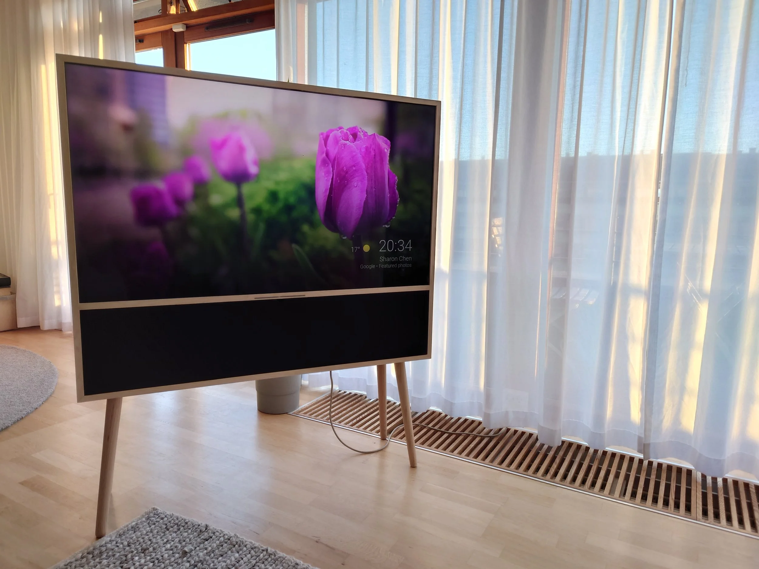 A large television displaying a close-up image of purple tulips, placed in a bright room with wooden floors, white curtains, and sunlight coming through the windows.