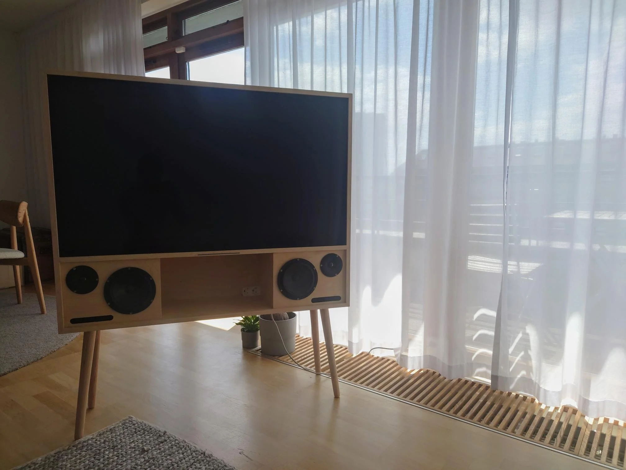 Modern flat-screen television with wooden speakers below, set on angled wooden legs in a bright room with sheer white curtains and a small potted plant on a wooden ledge.