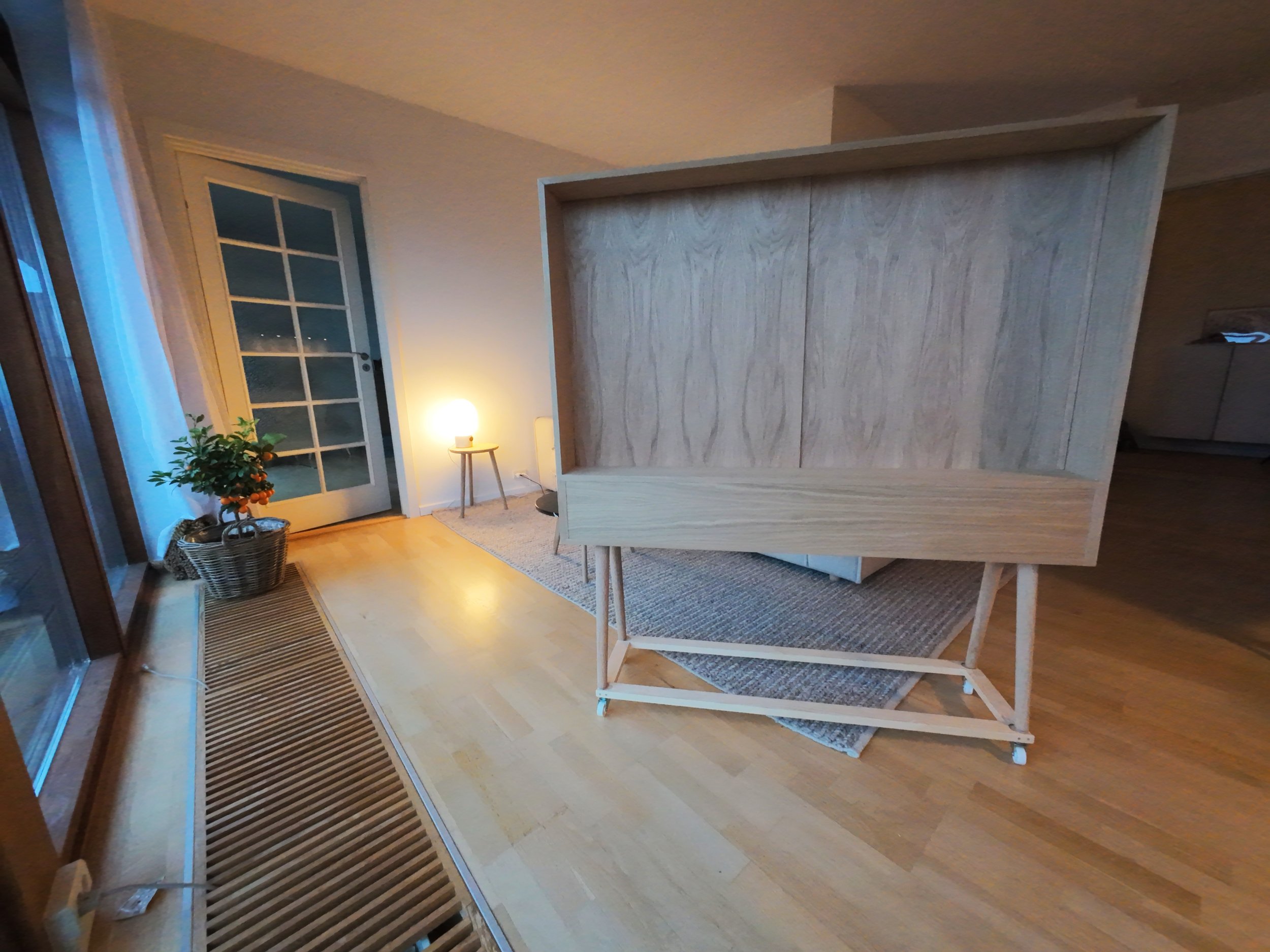 Interior with wooden furniture and warm lighting, a door with multiple glass panes, a potted plant, and a small side table with a lit lamp.