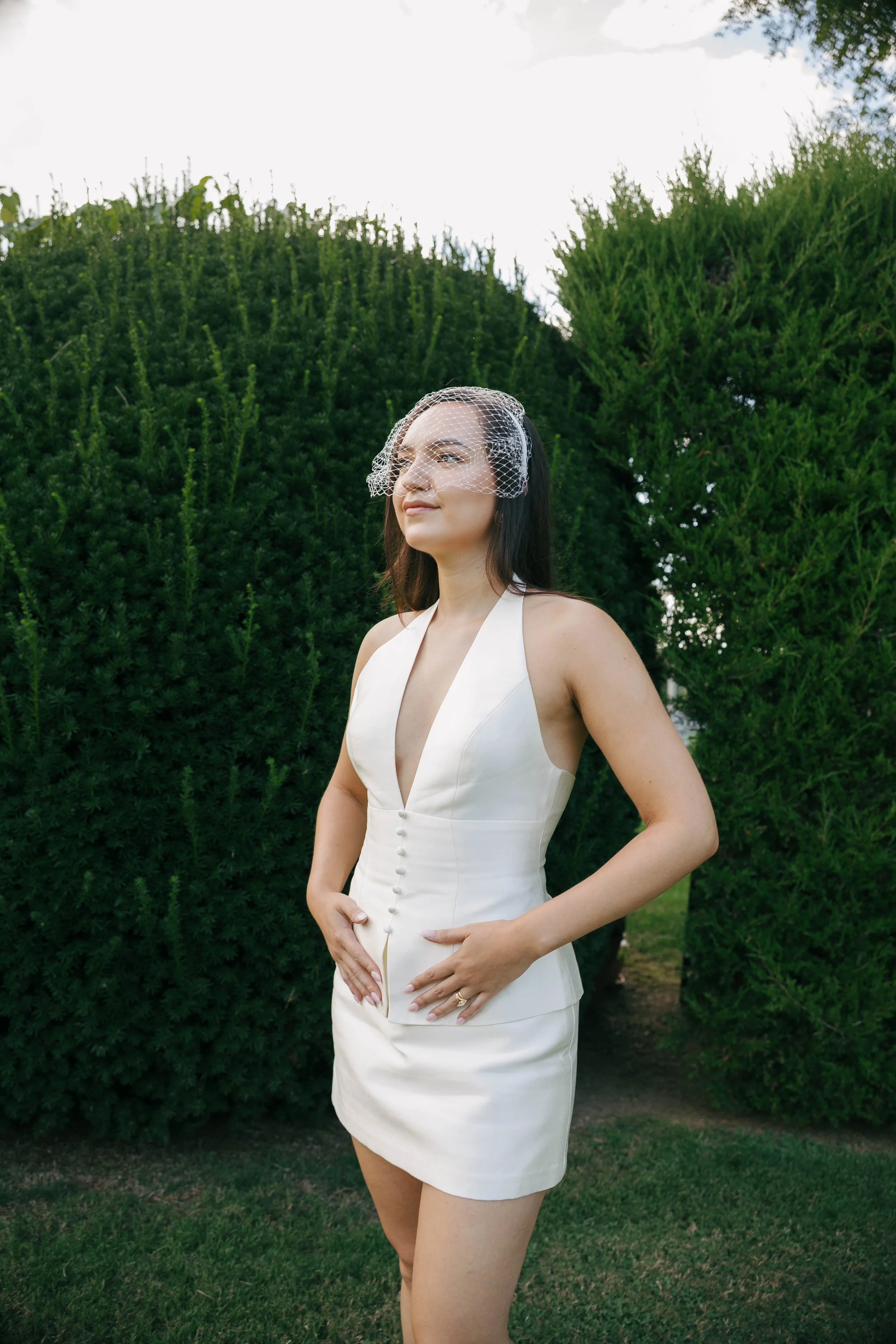 A woman in a white dress standing outdoors with green bushes in the background, wearing a birdcage veil.