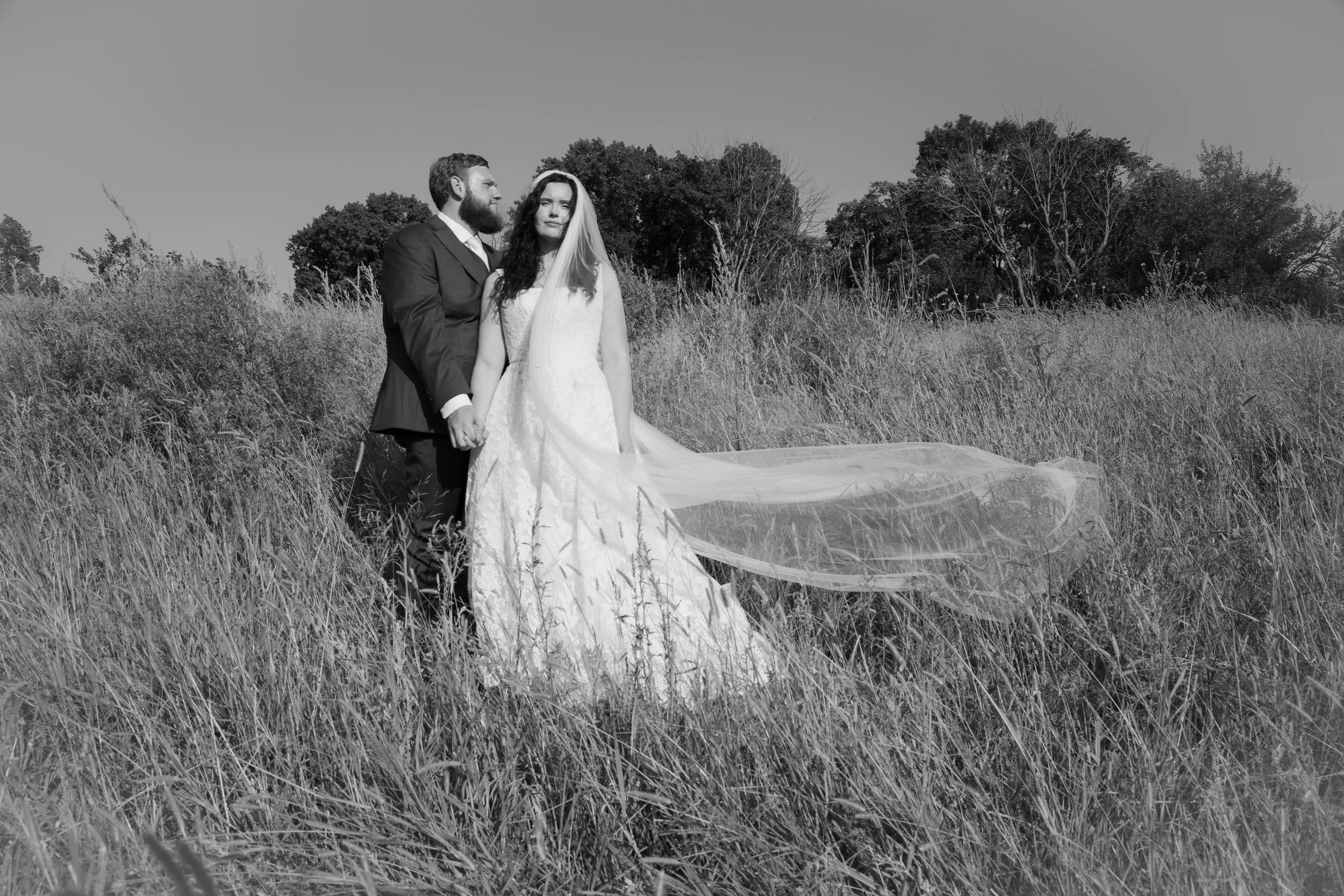 A black and white photo of a bride and groom holding hands in a grassy field, with trees in the background.