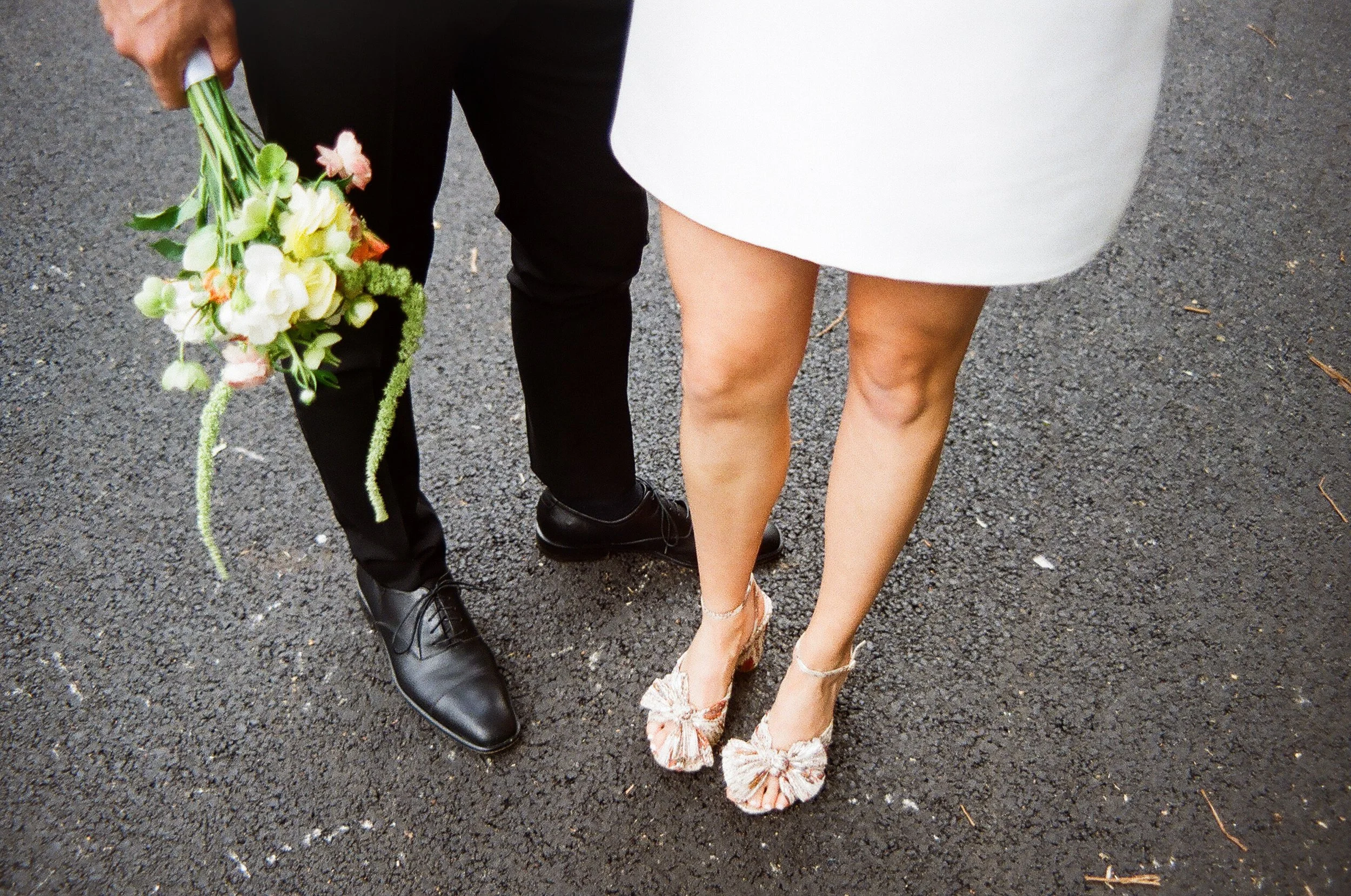 Close-up of a bride and groom standing on a rough asphalt surface, with the groom holding a bouquet of white and pink flowers.