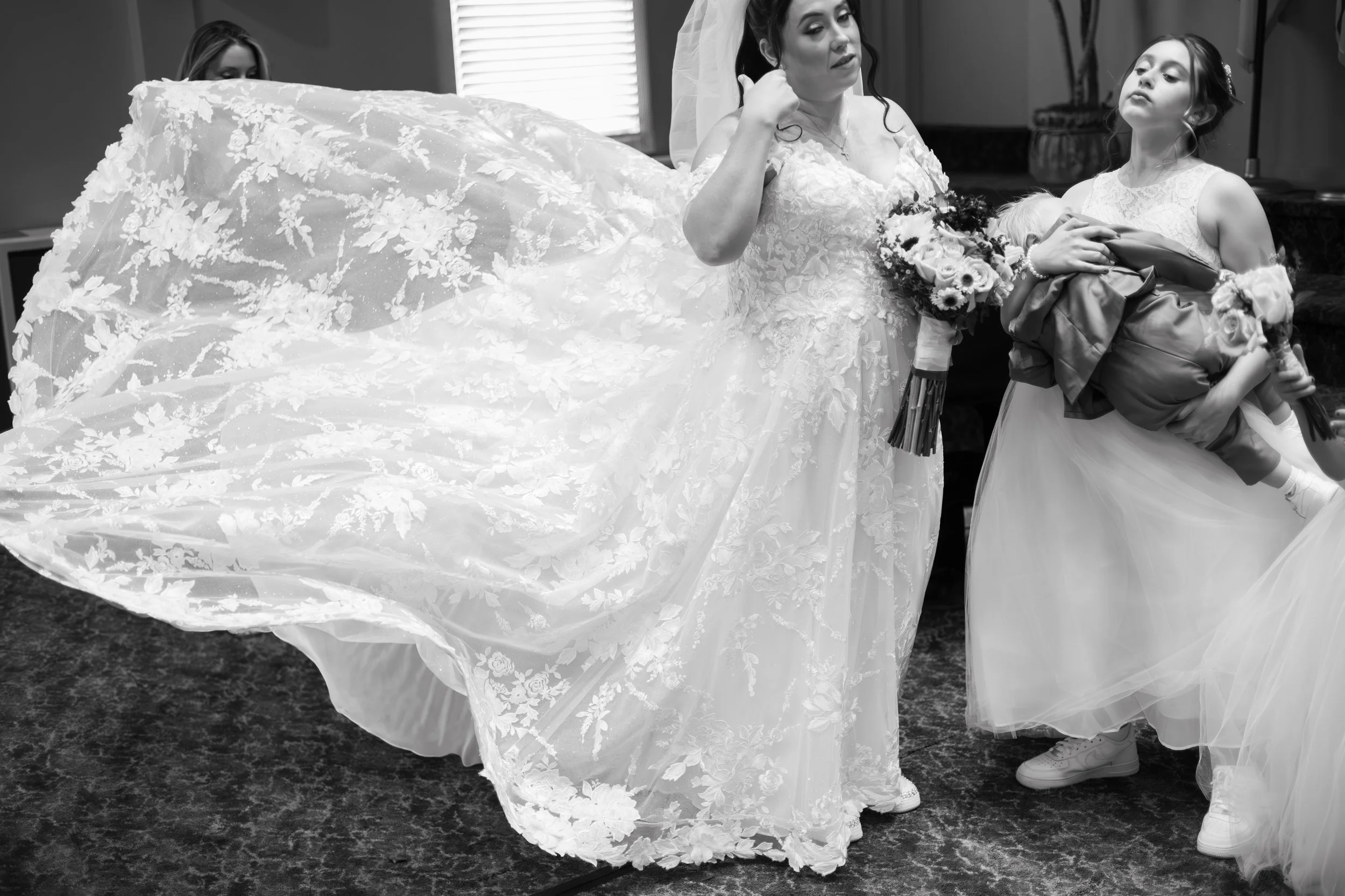 Black and white photo of a bride with a bouquet, a young girl holding a bouquet and a woman holding a child, possibly during a wedding event.