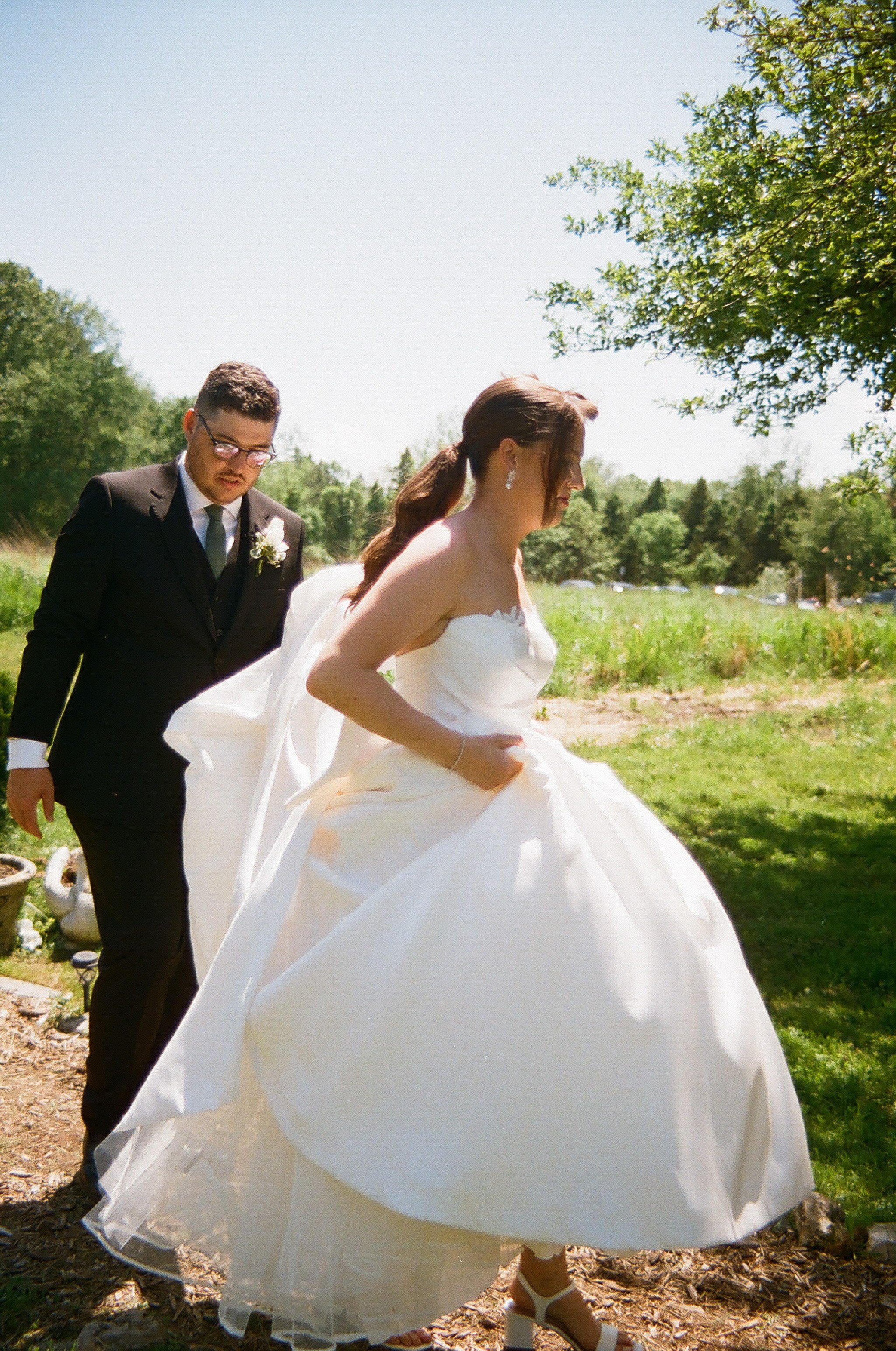 A bride and groom outdoors, with the bride lifting her wedding dress slightly as she walks, under a tree on a sunny day.