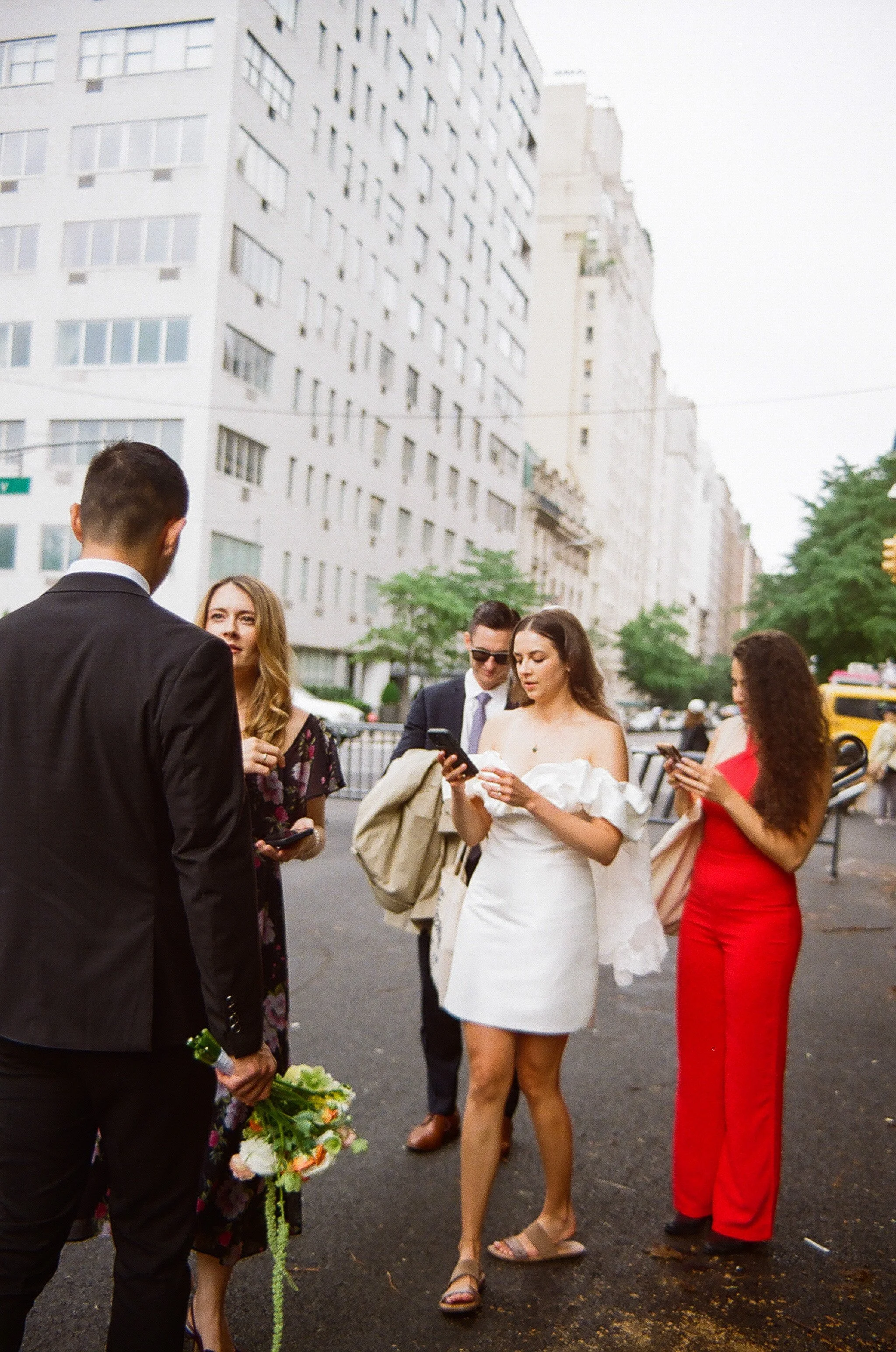 Group of people standing outdoors on a city street, using their smartphones and holding flowers, with tall white buildings in the background.