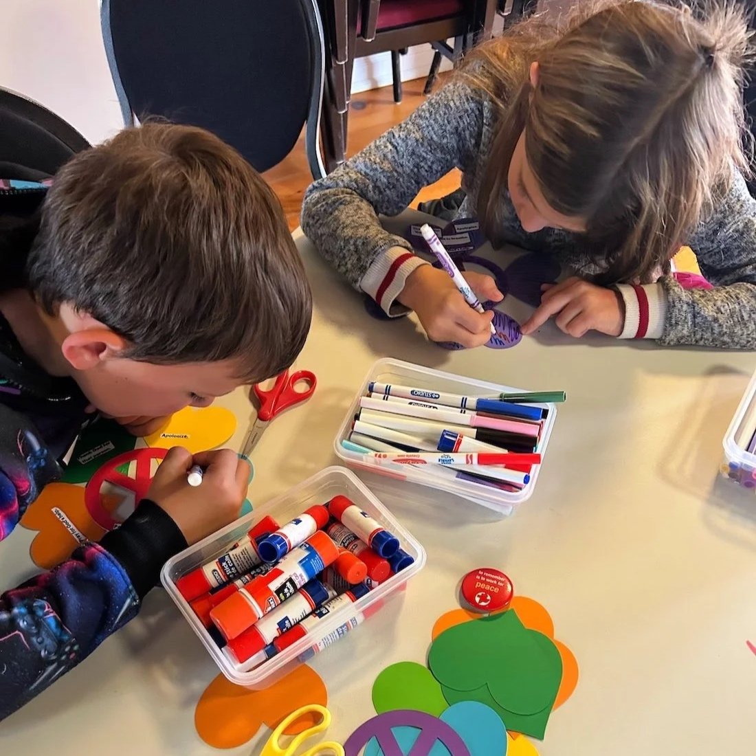 Two children in children's ministry working on crafting projects at a table with markers, glue sticks, scissors, and paper cutouts.