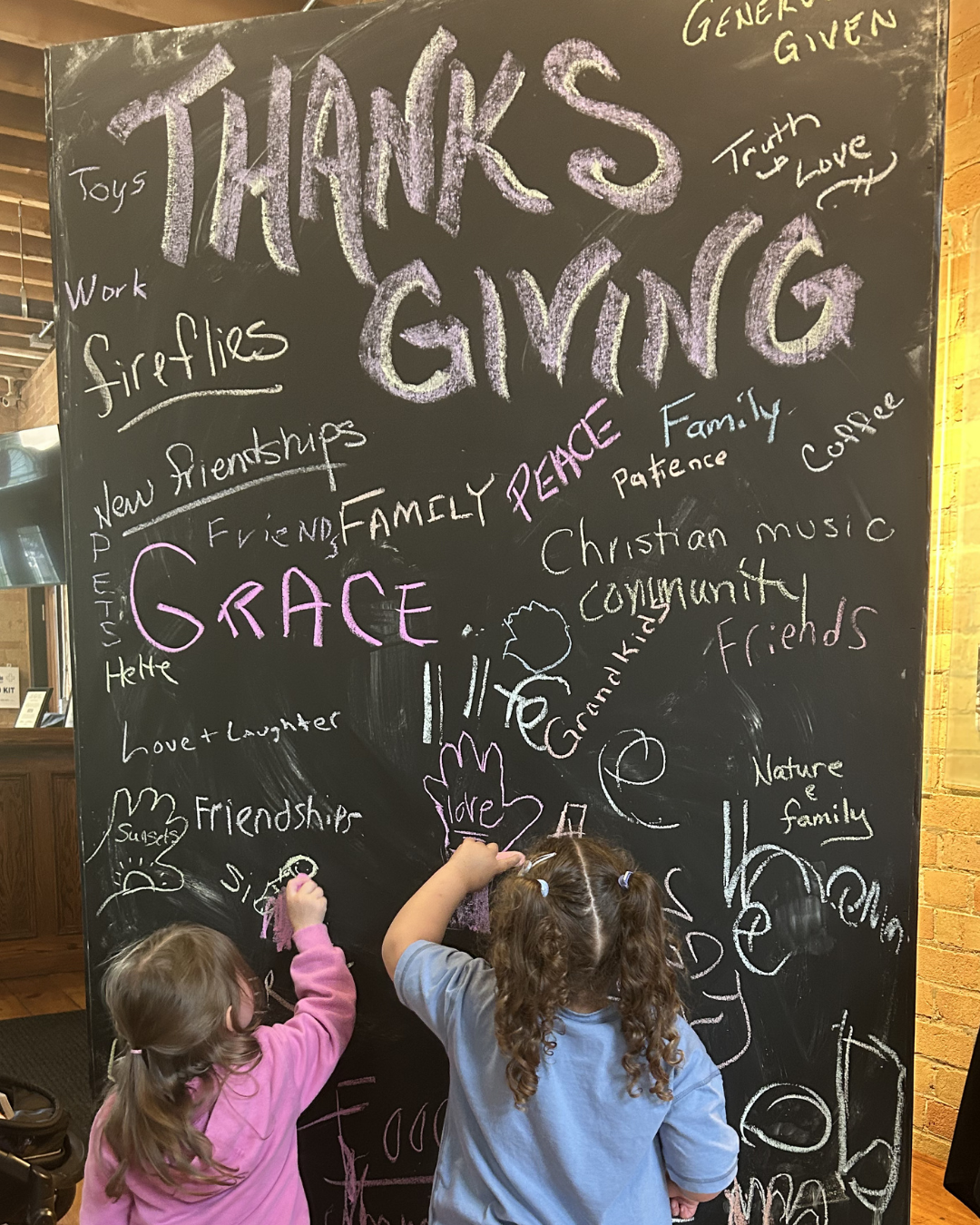 Two young girls with curly hair, one wearing a pink jacket and the other in a gray shirt, drawing on a large black chalkboard with colorful chalk. The chalkboard displays an assortment of words and phrases related to gratitude, including 'THANKS GIVING,' 'GRACE,' 'PEACE,' 'family,' 'friendship,' and 'love,' towards the top.