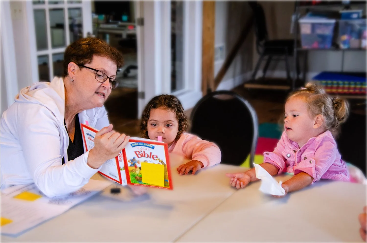 An adult woman reading a toddler Bible to two young girls at a table in a children's ministry class.
