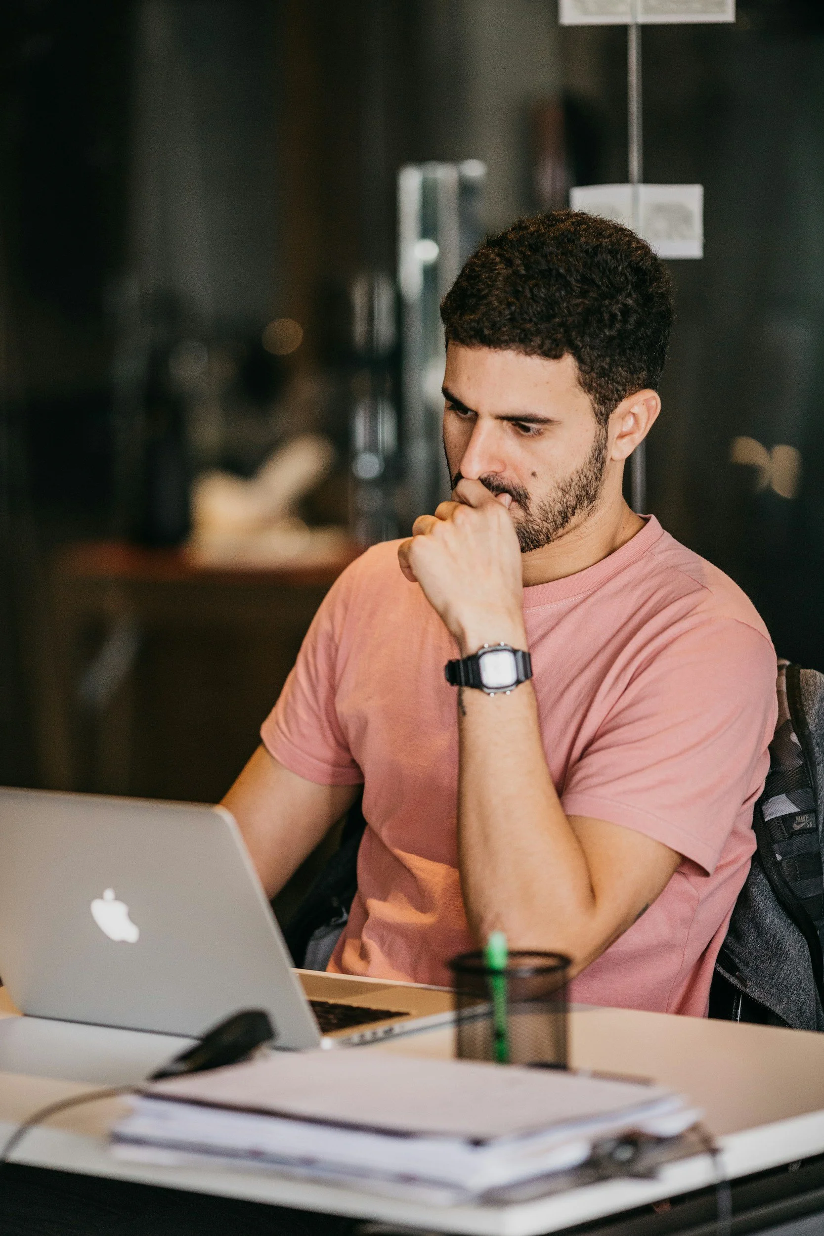 A man with dark, curly hair and a beard is sitting at a desk, resting his chin on his right hand and looking at a laptop with an Apple logo. He is wearing a pink T-shirt and a black watch. There are papers, a pen, and a cup with a straw on the desk. The background is dark and blurred.