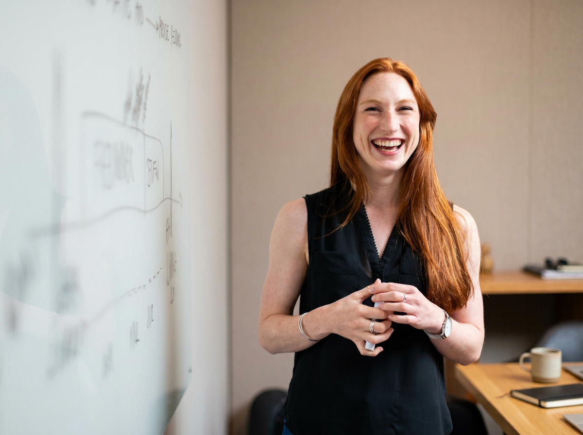 Young woman with long red hair laughing in a meeting room, standing beside a whiteboard with writing on it.
