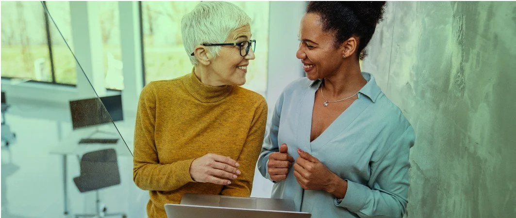 Two women smiling and having a conversation in an office setting.