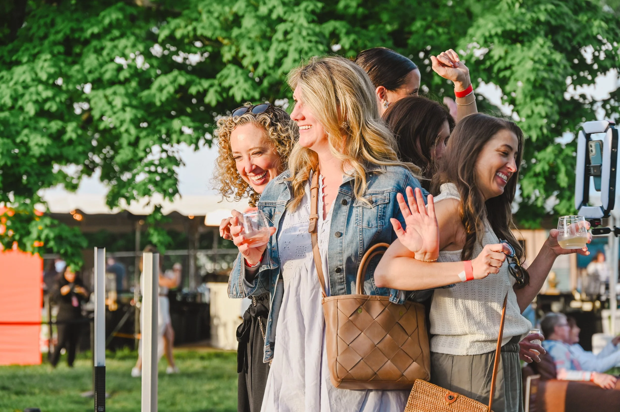 Group of smiling women and one man attending a social event, some holding glasses of wine, standing under string lights in an indoor venue.