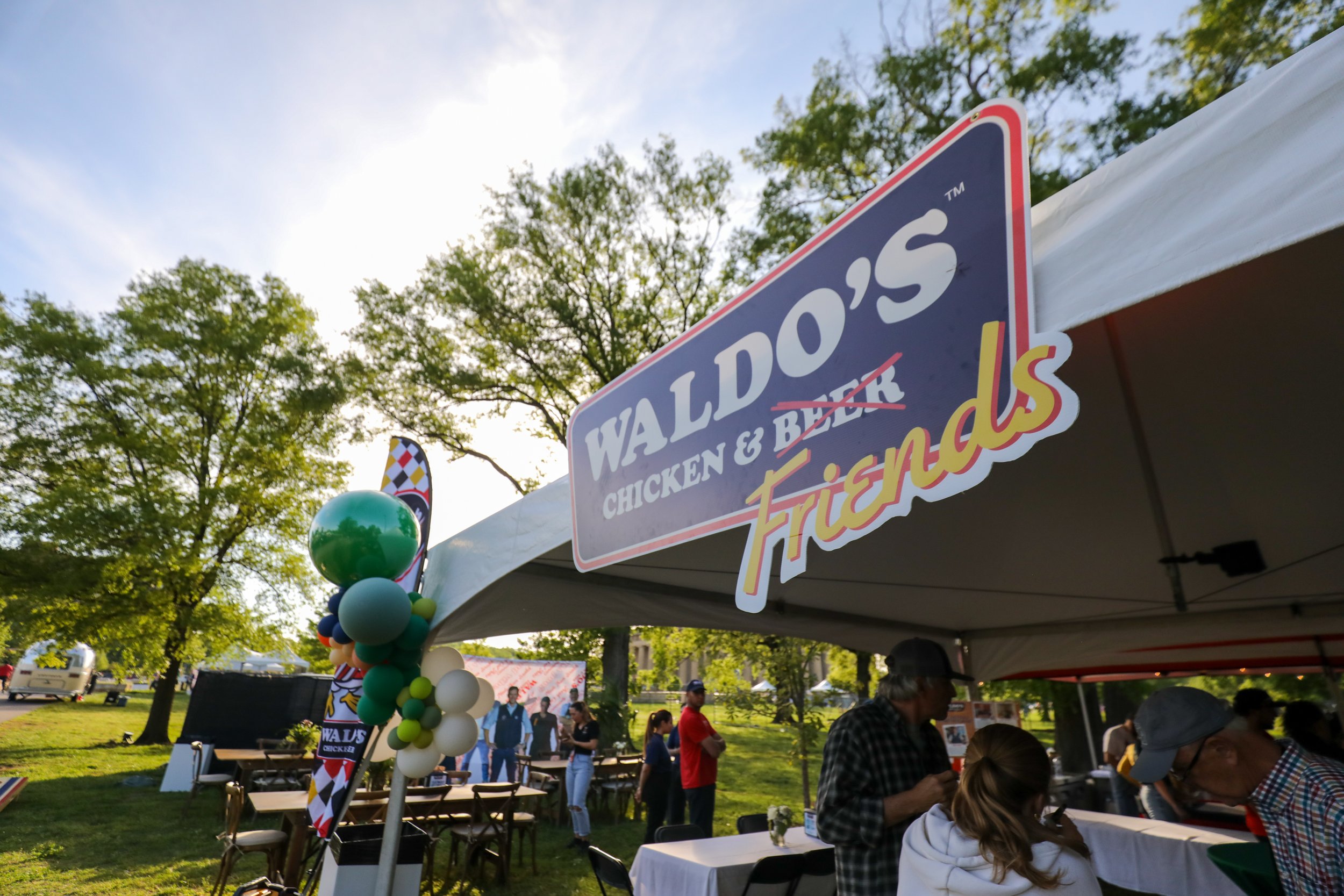 A man wearing a cowboy hat and a colorful apron cooks food with flames on an outdoor cooking station during evening, with a group of people watching and taking photos in the background.