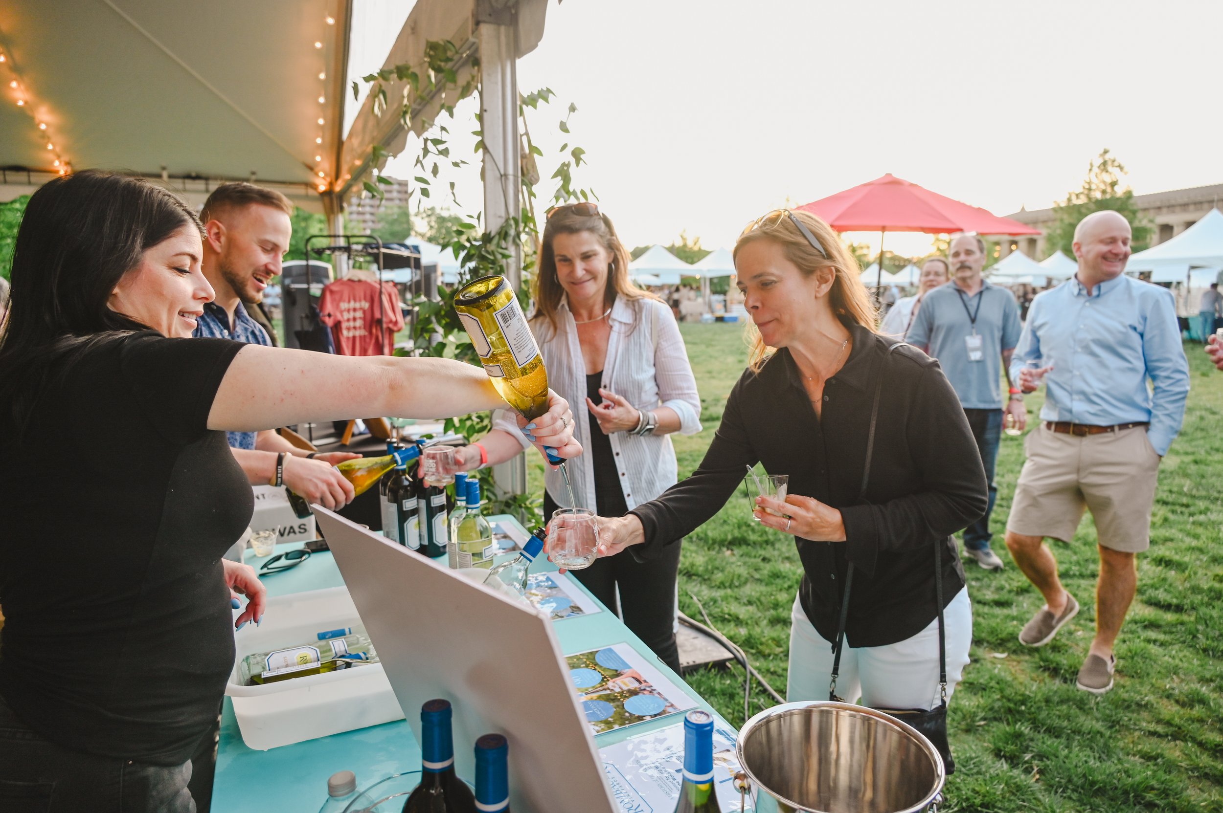 People seated at a long outdoor table enjoying a meal, with a waiter pouring a drink, and tents and buildings in the background.