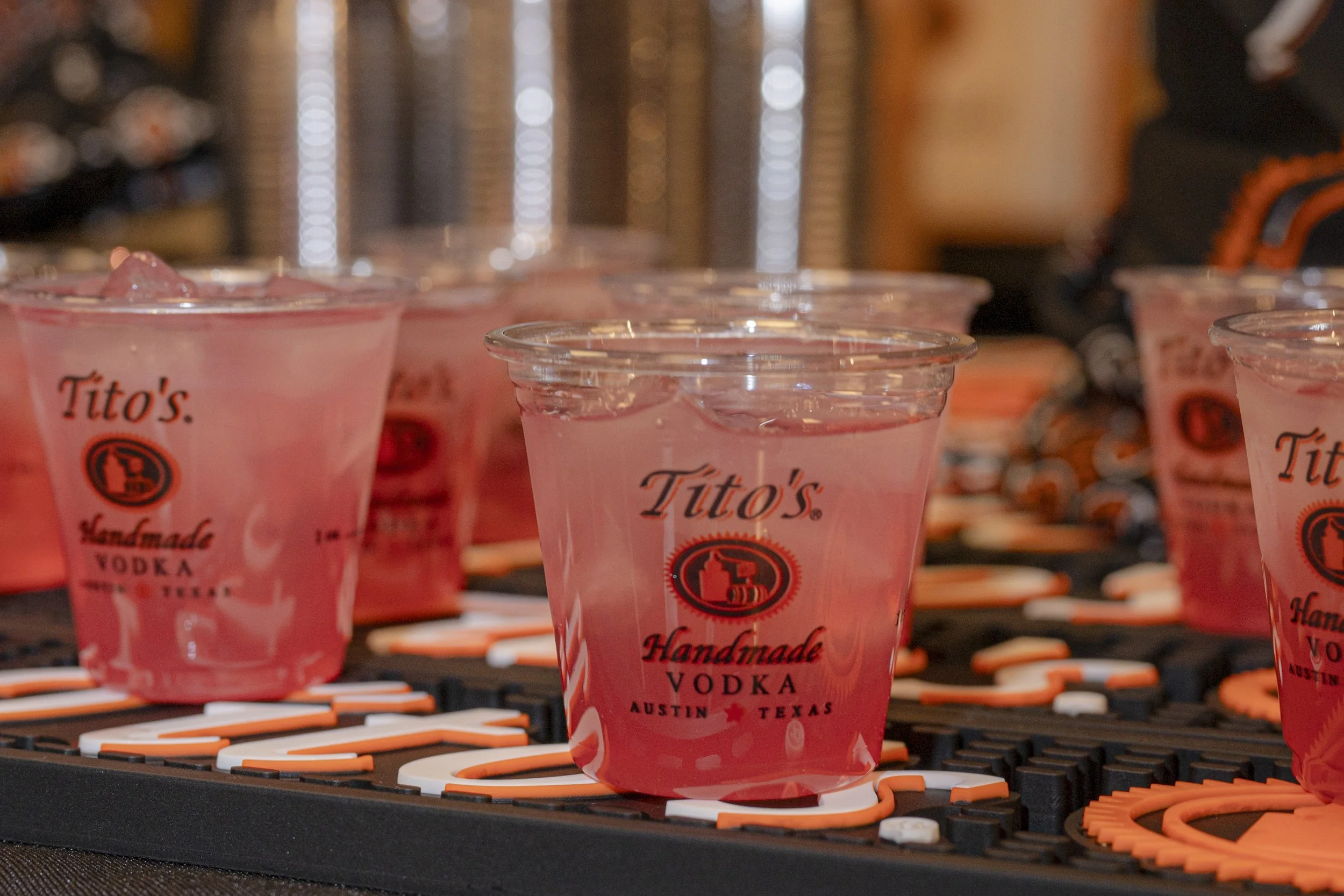Clear plastic cups filled with pink-colored vodka drinks on a black tray with orange accents, branded with 'Tito's Handmade Vodka' and 'Austin Texas'.