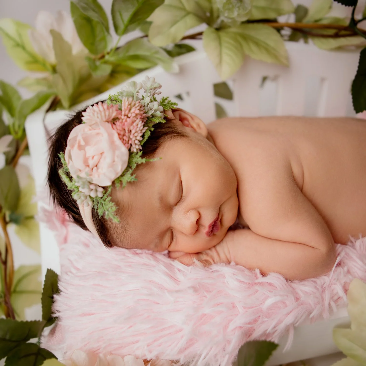 Newborn baby sleeping with a floral headband, lying on a soft pink blanket, surrounded by greenery.