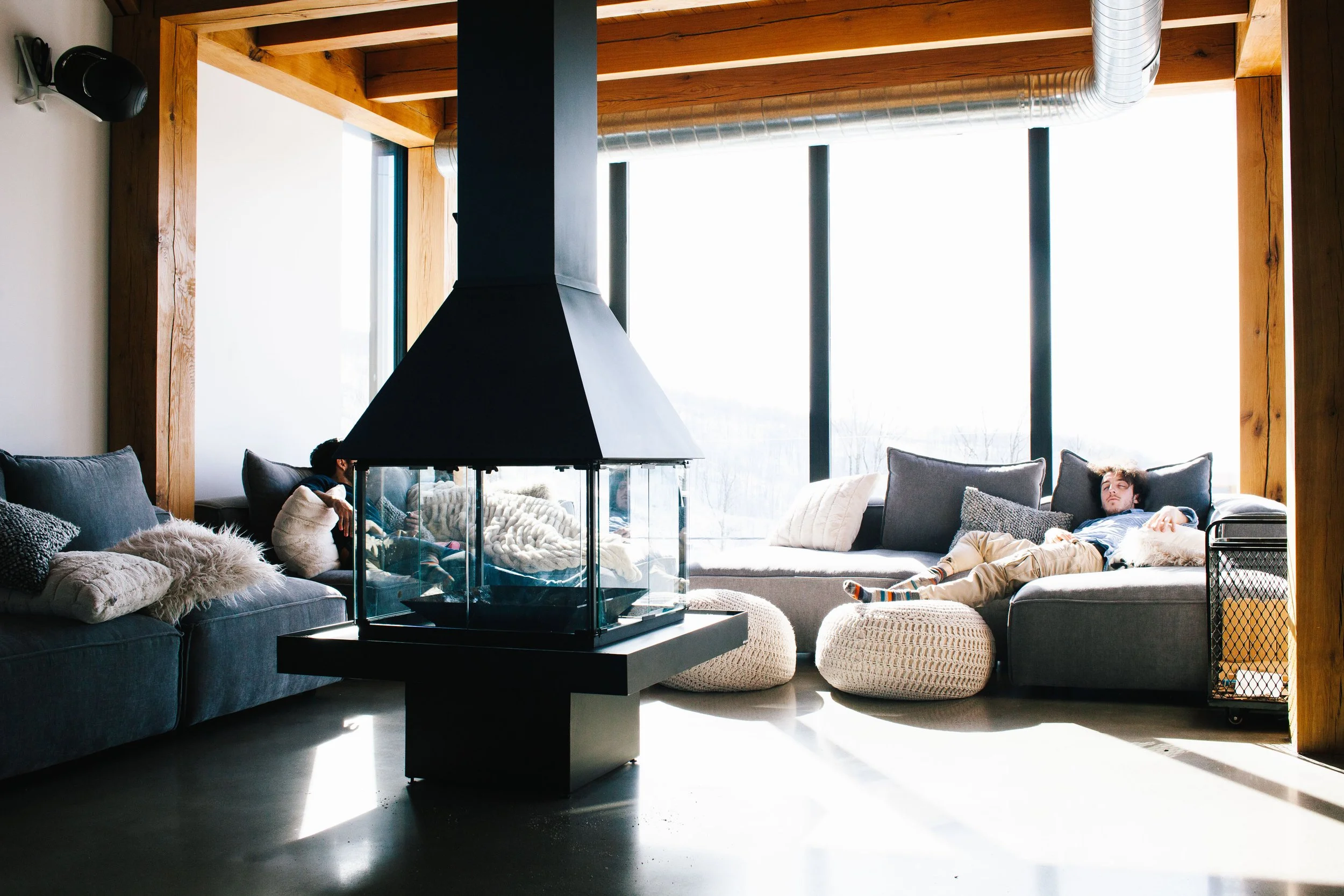 Salon moderne avec grande fenêtre, sofas en tissu gris, coussins, poufs ronds en tricot, cheminée en verre, bois au plafond, personne allongée sur un sofa, lumière naturelle.