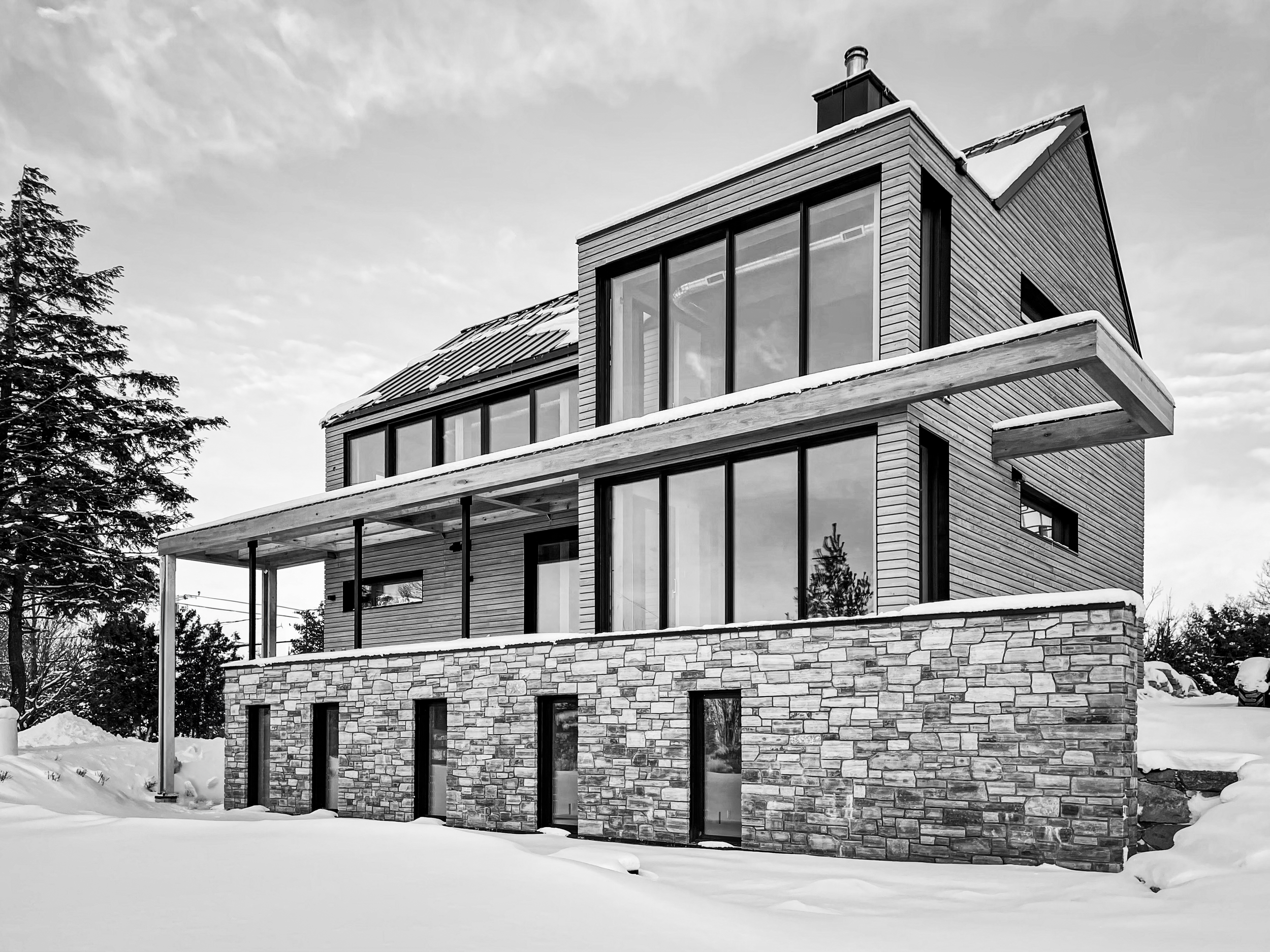 Maison moderne en bois et pierre avec plusieurs étages, grandes fenêtres, balcon en dessous et environnement enneigé.