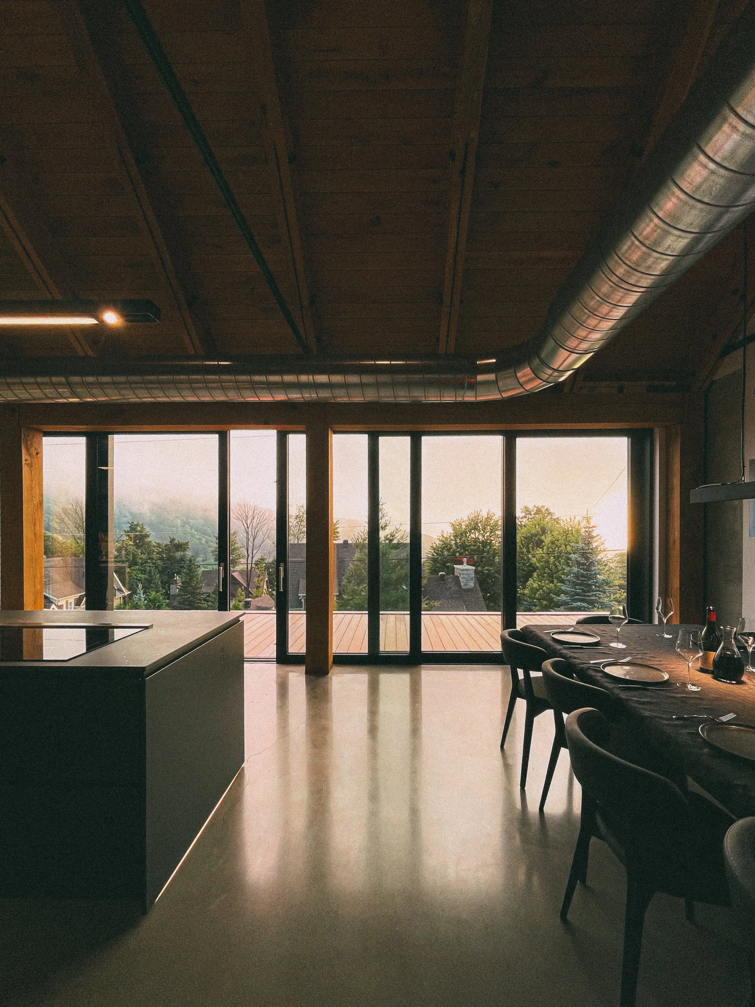 Intérieur d'une salle à manger moderne avec grande baie vitrée donnant sur un paysage avec des arbres, et une table dressée avec des assiettes, des verres et une bouteille de vin.