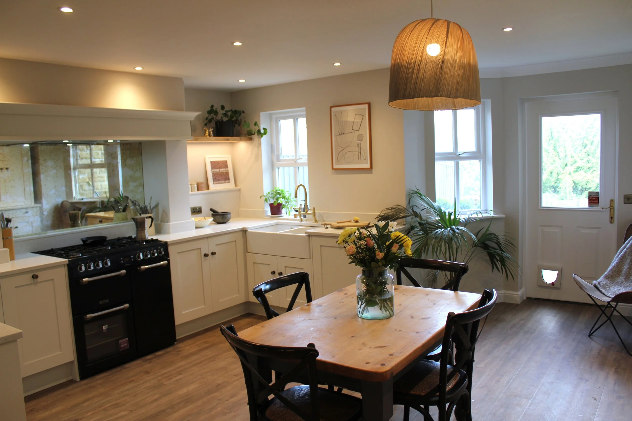 A cozy kitchen with white cabinets, a black stove, a wooden dining table with black chairs, potted plants, framed artwork, and large windows letting in natural light.