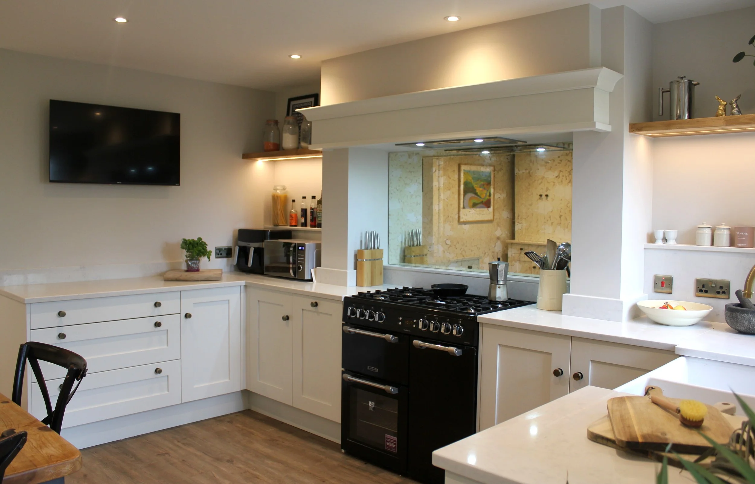 Modern kitchen with white cabinets, a black gas stove, and a built-in range hood. There is a flat-screen TV mounted on the wall, open shelves with jars and decorative items, and a small plant on the counter. The kitchen has a light, neutral color pal