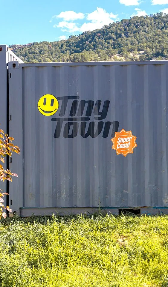 Shipping container with hand-lettered "Tiny Town" wordmark and "Super Cozy" badge graphic

A weathered shipping container at Tiny Town carries a hand-drawn wordmark, smiley icon, and "Super Cozy" badge, part of the community's playful environmental b